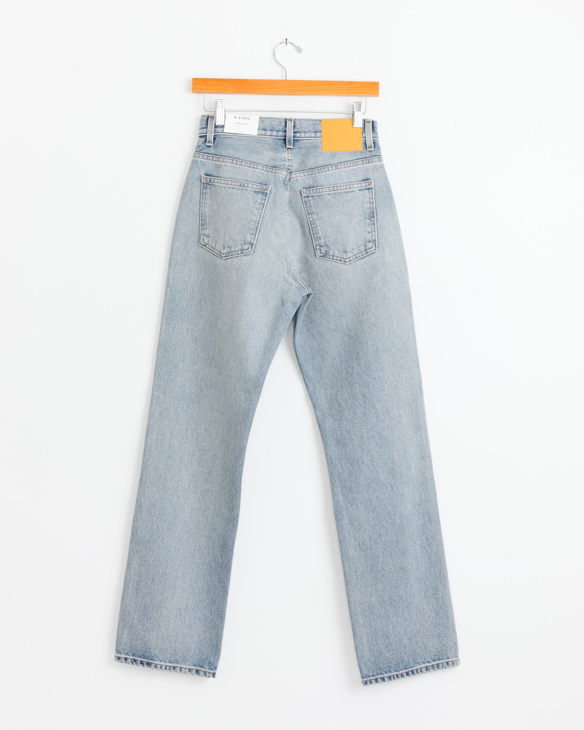 The Mira Jean in Ed Wash by B Sides hangs from a wooden hanger against a white background, displaying the back with two pockets and a tan leather patch on the waistband.