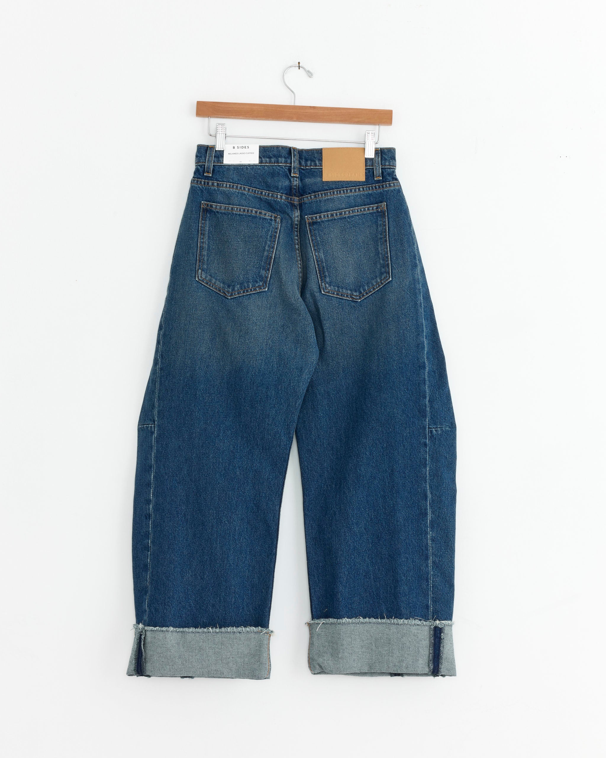 The B Sides Relaxed Cuff Lasso in Cate Wash are blue heavyweight cotton denim jeans with wide legs, large cuffs, two back pockets, and a brown leather waistband patch, shown on a wooden hanger against a white background.