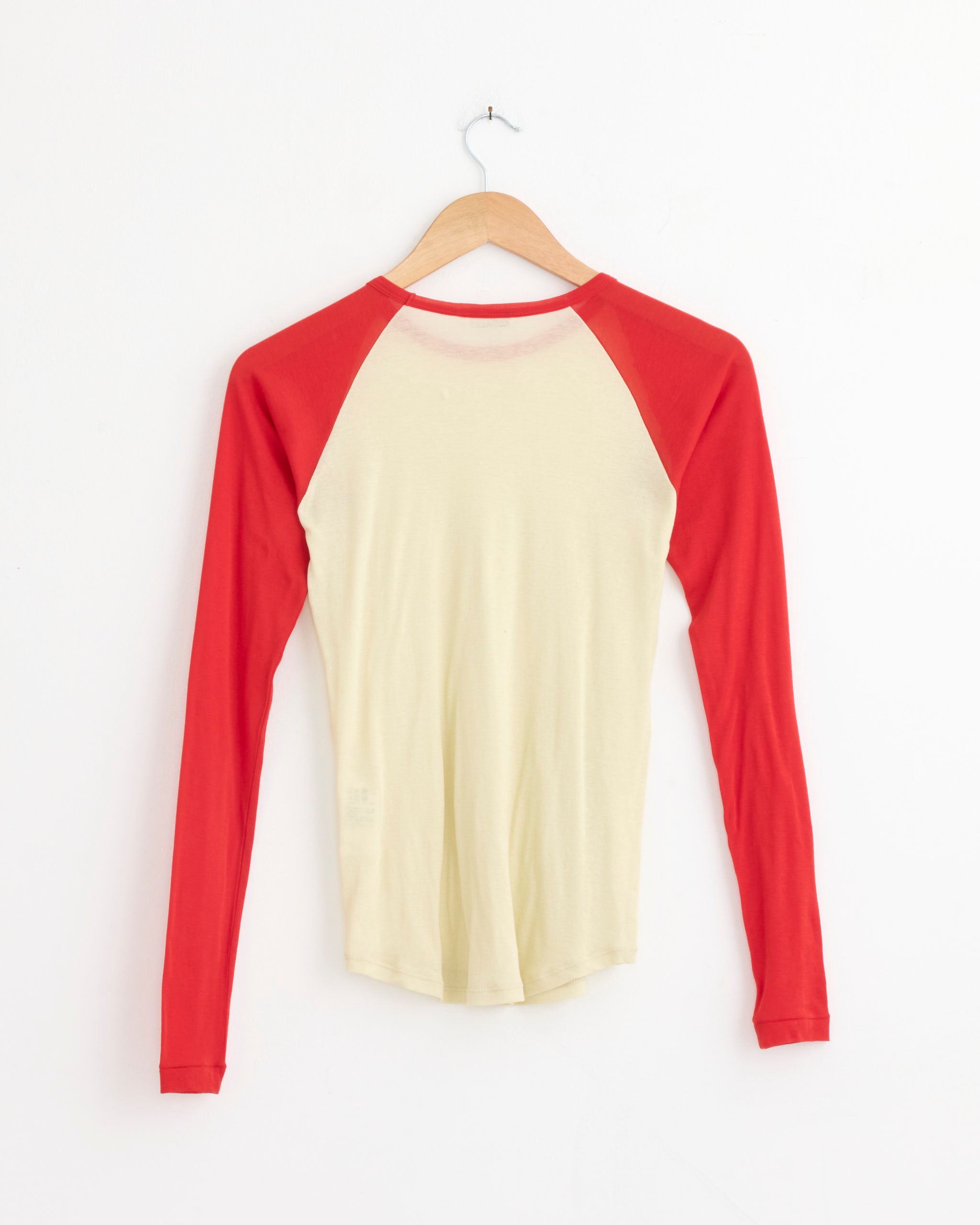 The Auralee Super High Gauge Sheer Pullover in Ivory/Red features long raglan sleeves and extra-long staple cotton, shown on a wooden hanger with the back facing outward against a white wall.