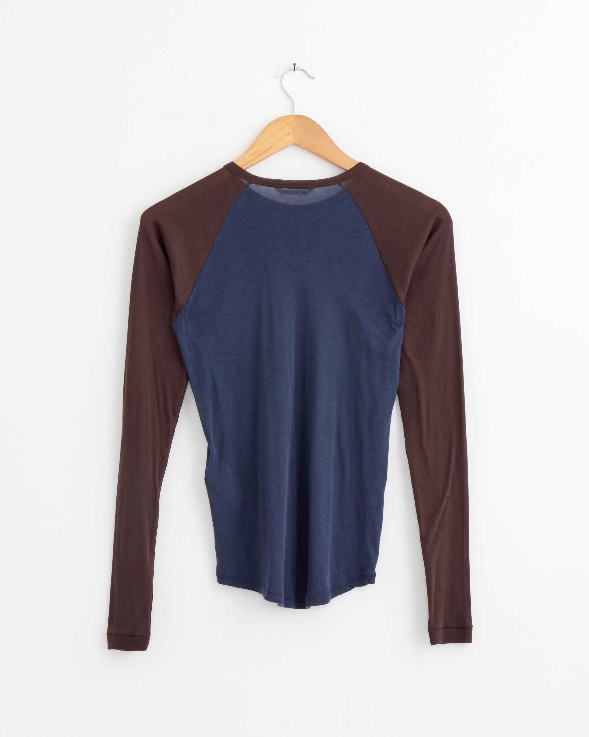 The Auralee Super High Gauge Sheer Pullover in Navy/Dark Brown, crafted in Japan from extra-long staple cotton, is shown hanging on a wooden hanger against a plain white wall.