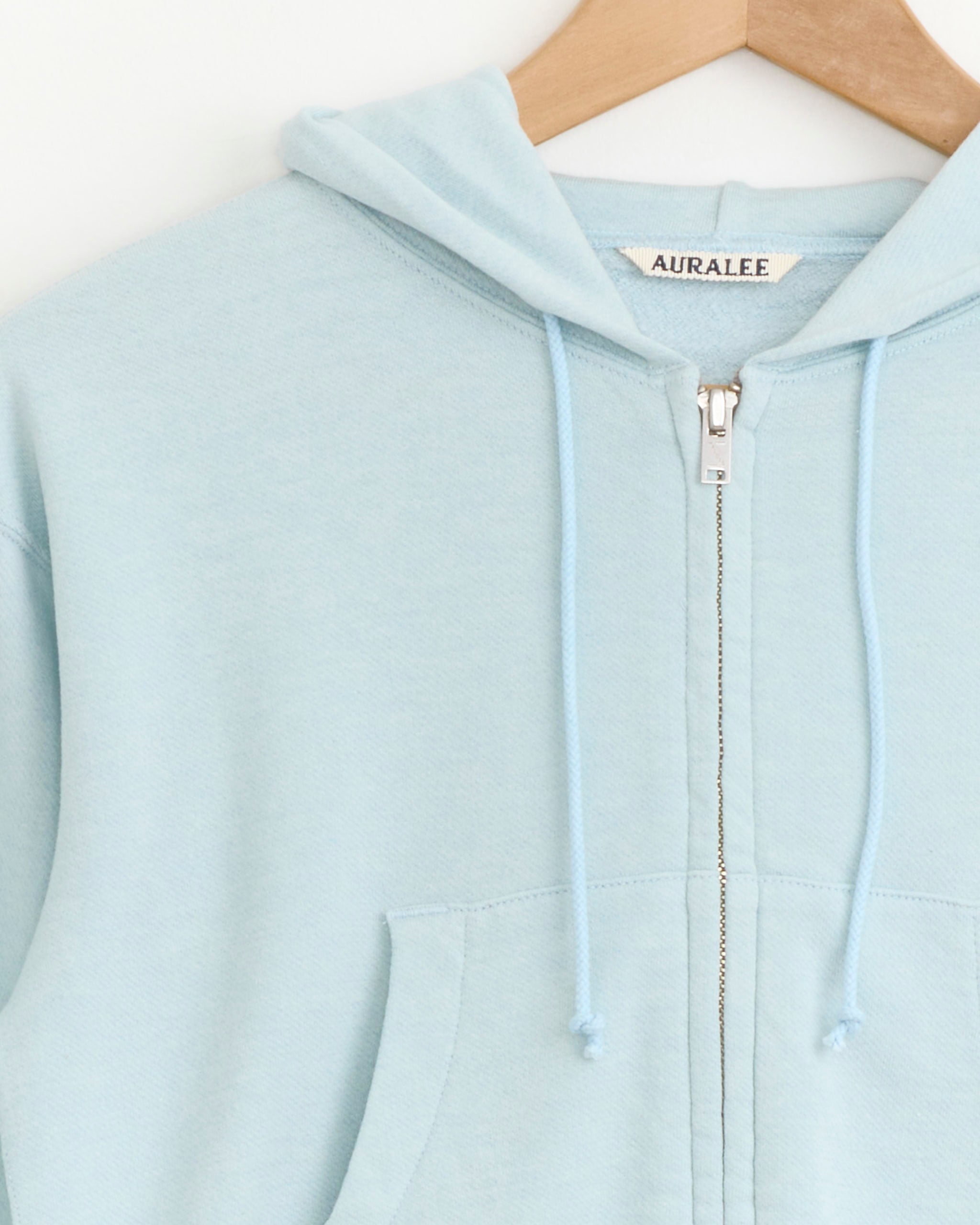 Close-up of the Auralee High Twist Light Sweat Zip Parka in Pale Blue, featuring a hood and drawstrings, hanging on a wooden hanger. The Auralee brand label is visible inside the collar.