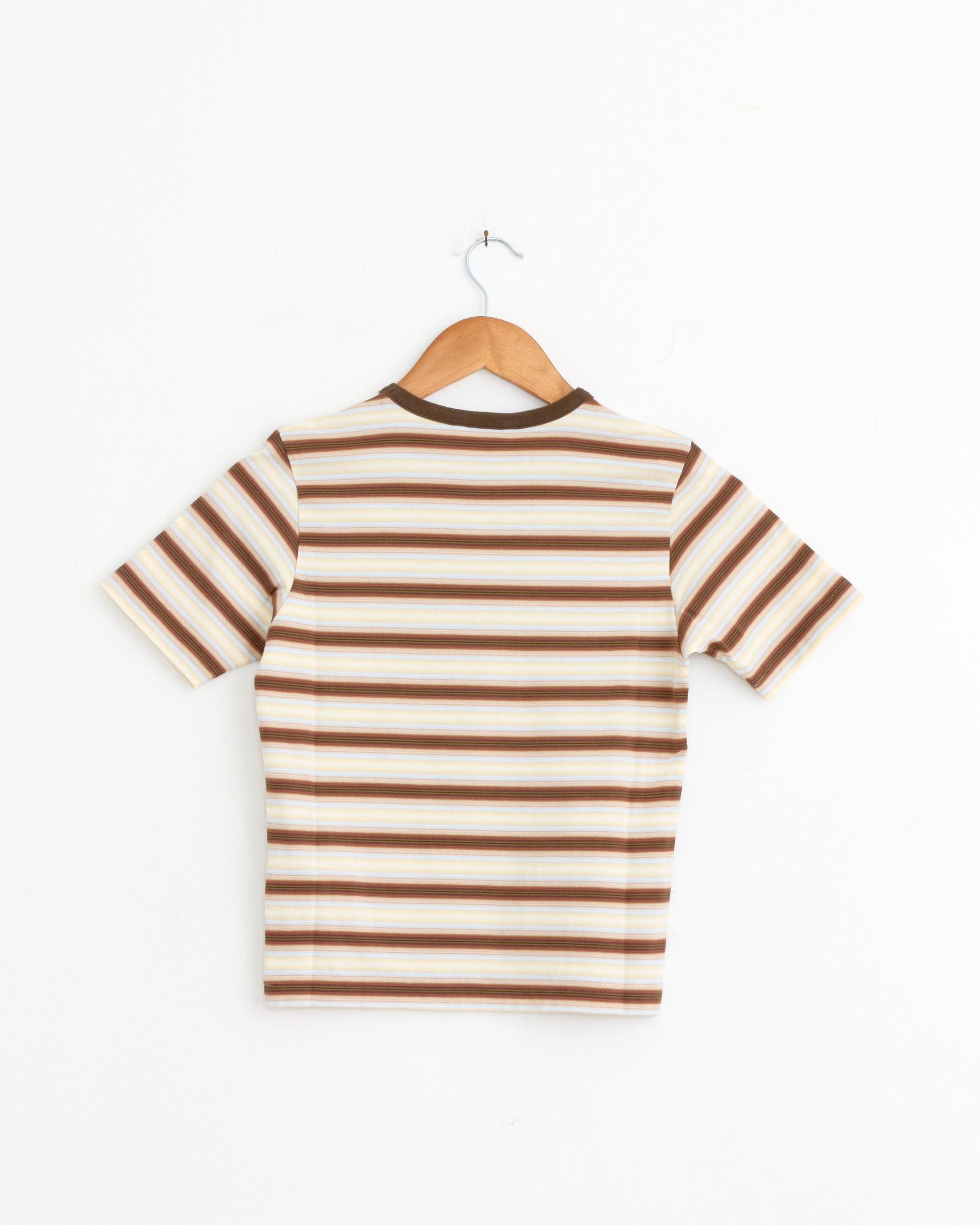 The Auralee Gradient Stripe Jersey Tee in Fade Brown Stripe, a short-sleeve cotton jersey with horizontal brown, beige, and cream stripes, is displayed from the back on a wooden hanger against a plain white wall.