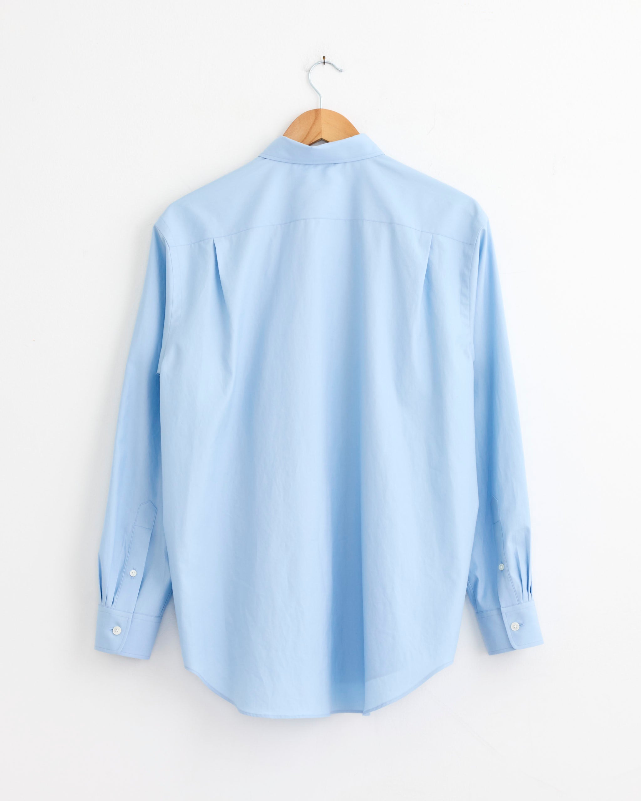 The Auralee Washed Finx Twill Shirt in Light Blue is shown hanging on a wooden hanger against a plain white wall, with its back facing forward.