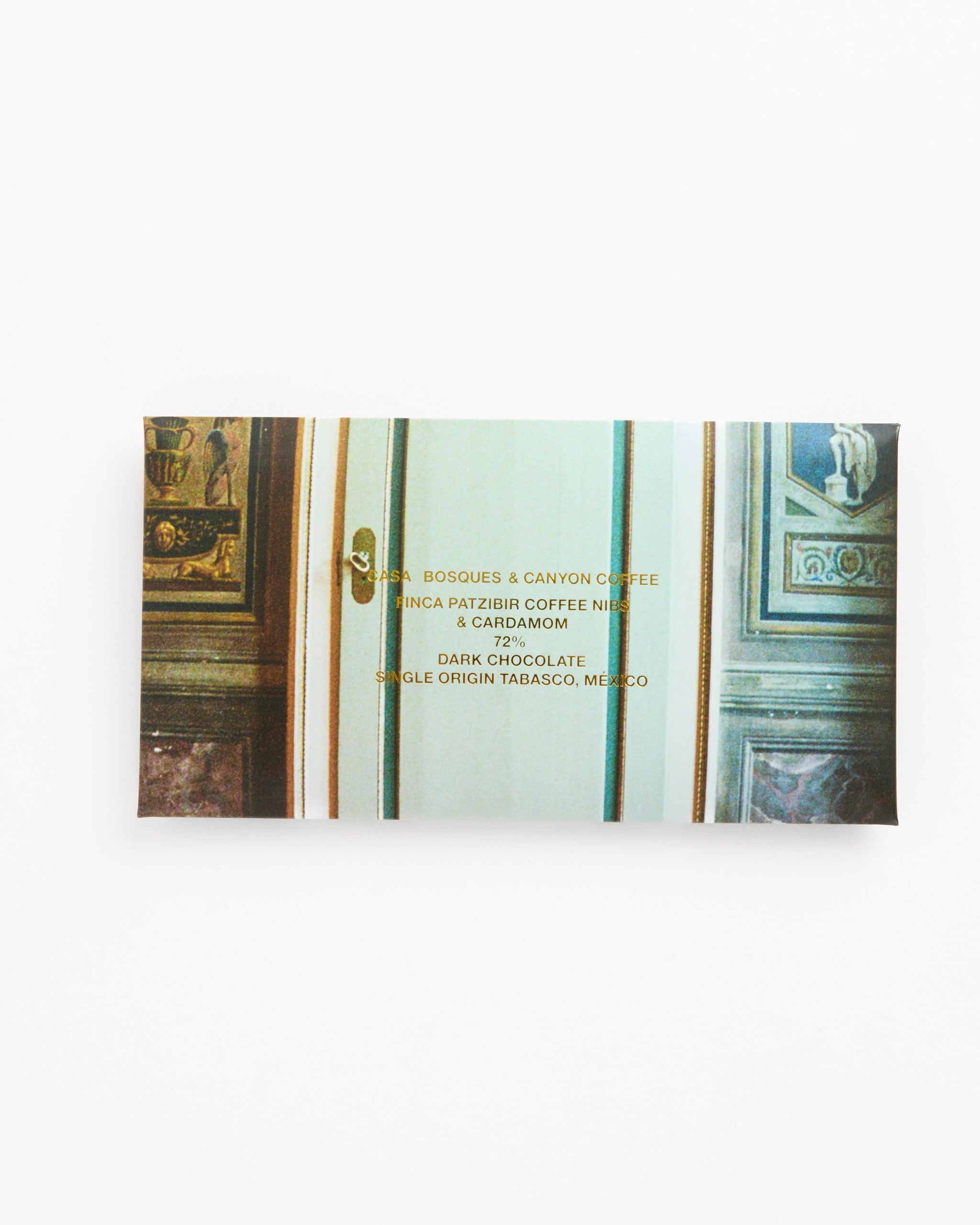 A Casa Bosques Canyon Coffee Chocolate bar featuring Guatemalan coffee beans and cardamom in single origin Tabasco chocolate, shown in decorative packaging with gold text against an ornate classical door.