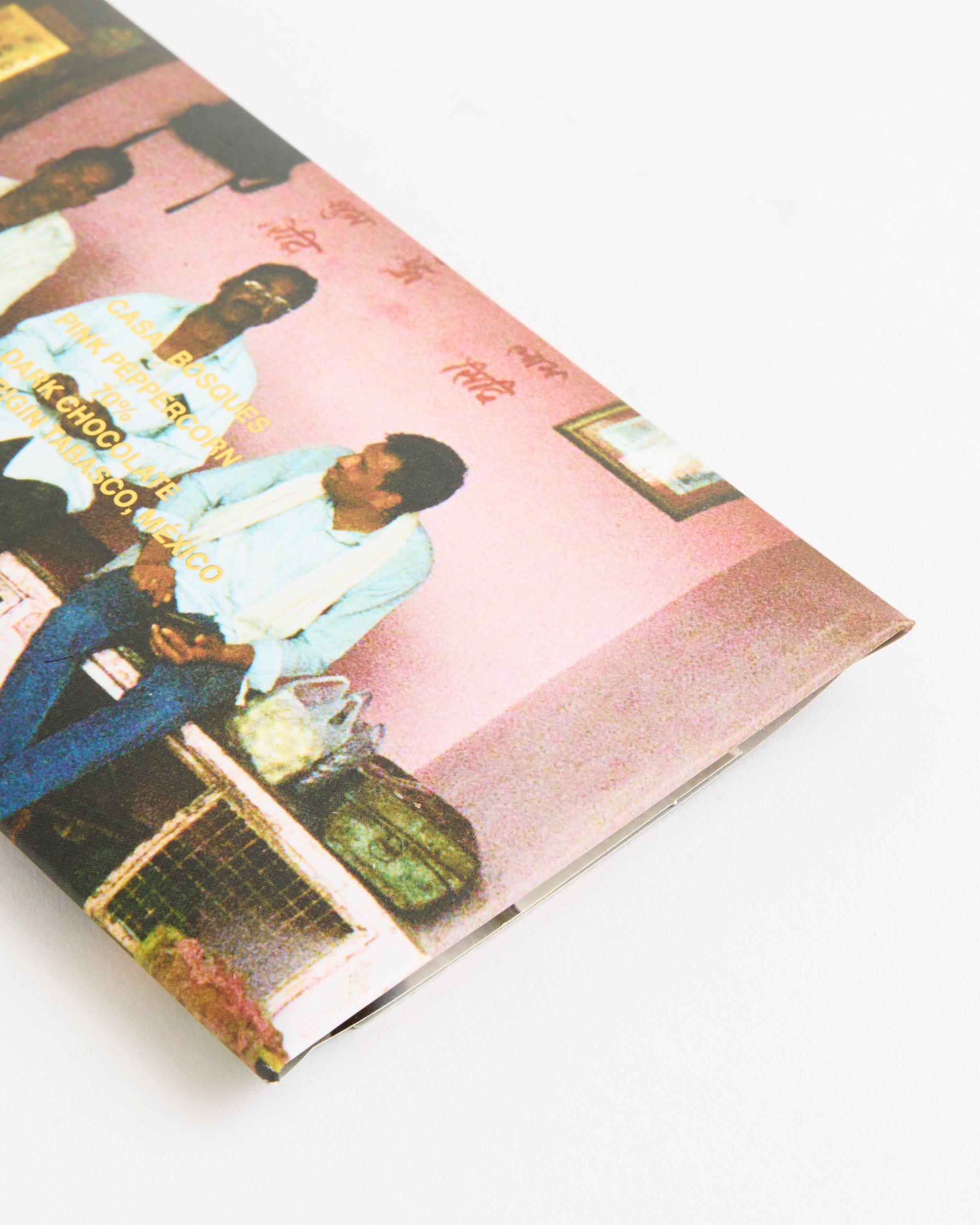 A close-up of the Casa Bosques Pink Peppercorn Chocolate book cover shows people in a pink room with yellow text, a green bag on a bench, and soft white fading edges, evoking the cozy ambiance of an artisan chocolate café.