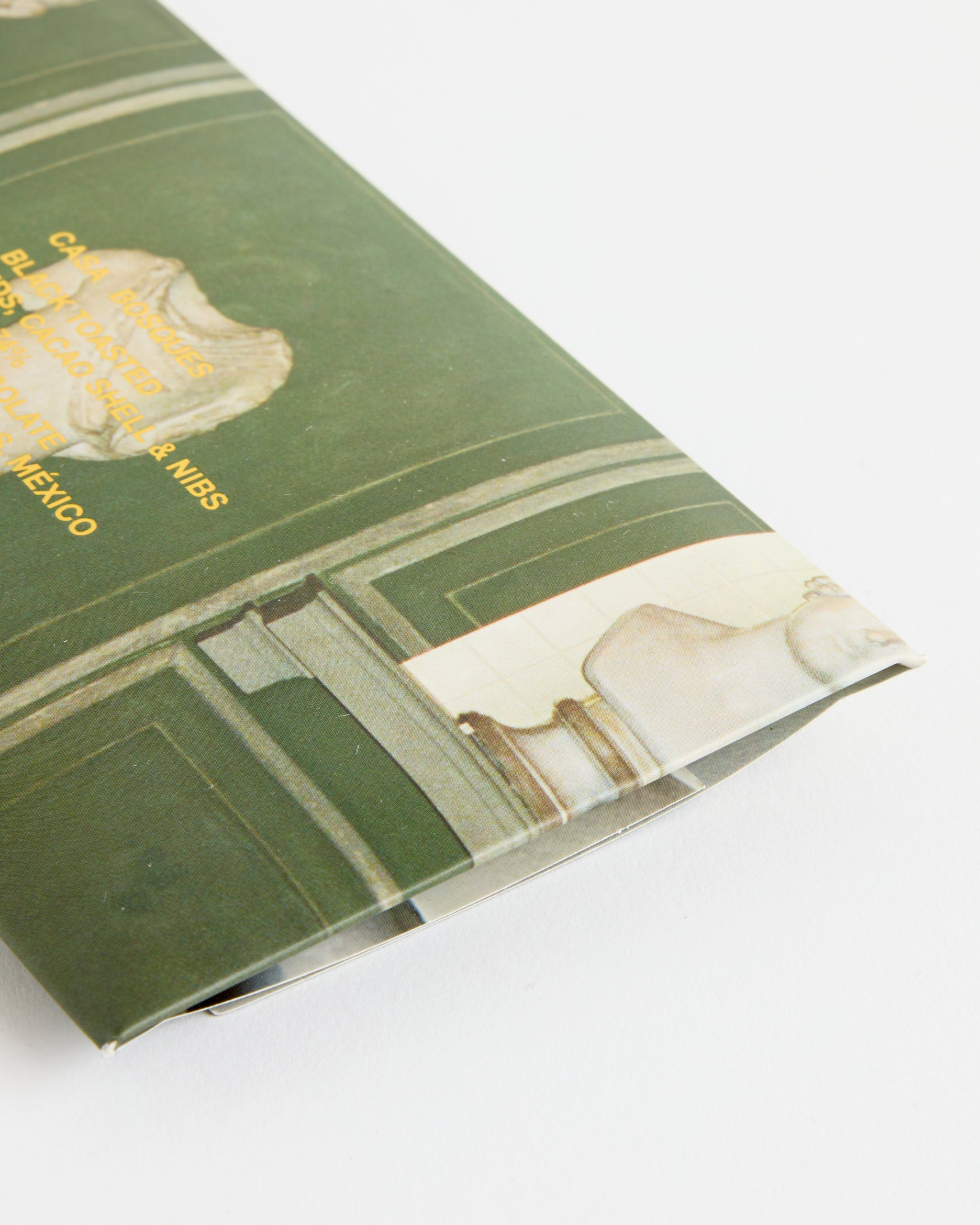 A close-up of a green book with gold text and classical statues on the cover, lying beside a Casa Bosques Black Toasted Sesame / Cacao Shell & Nibs Chocolate bar on a white surface.