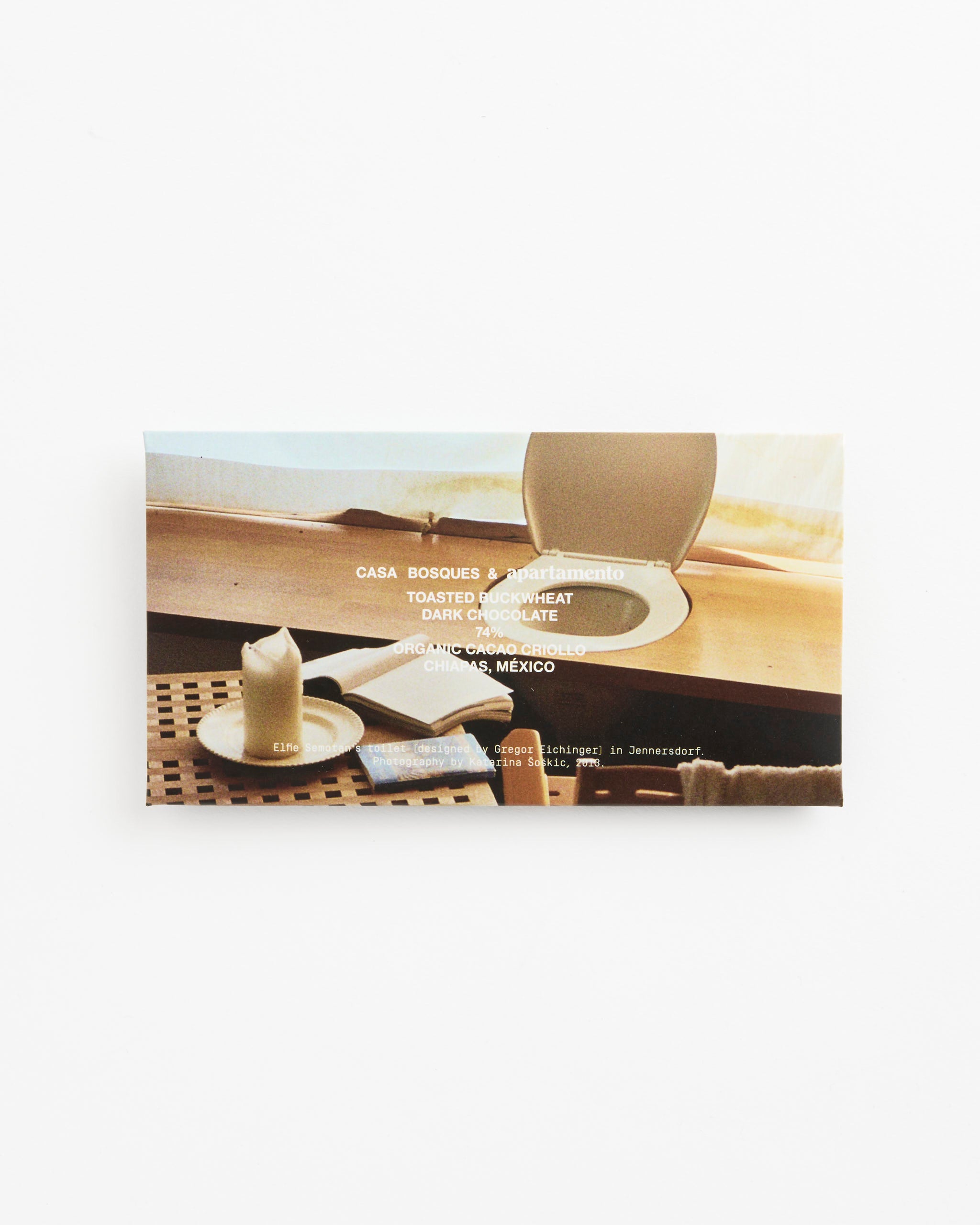 Casa Bosques Toasted Buckwheat x Apartamento Chocolate features single-origin dark chocolate with toasted buckwheat, in packaging illustrated with a cozy sunlit scene—a lit candle, cup, and round mirror on wood by a window.
