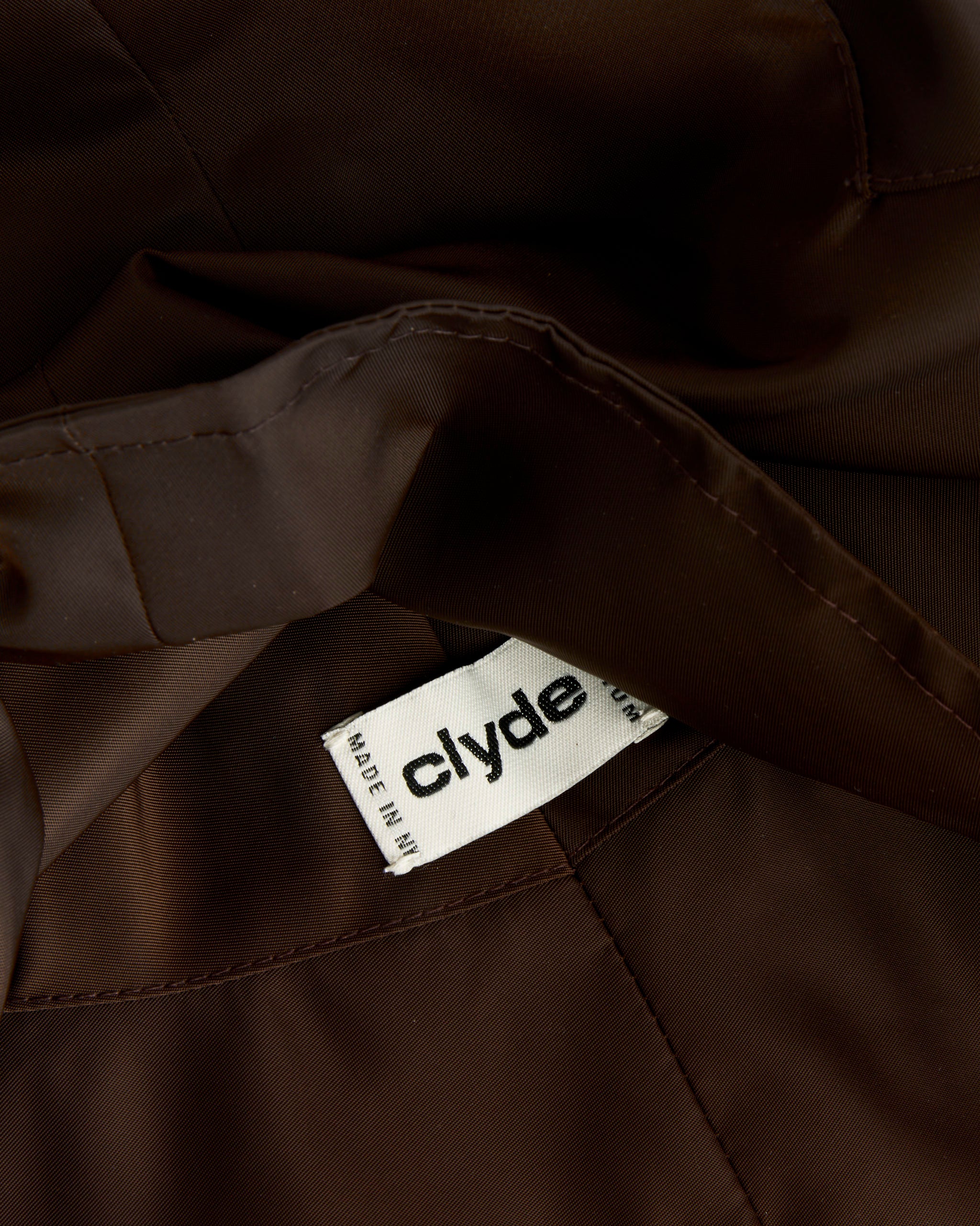A close-up of the Iona Hat in Espresso Flight Satin by Clyde shows a white fabric label with "clyde" in black text, partially visible and sewn into the seam for added sun protection.