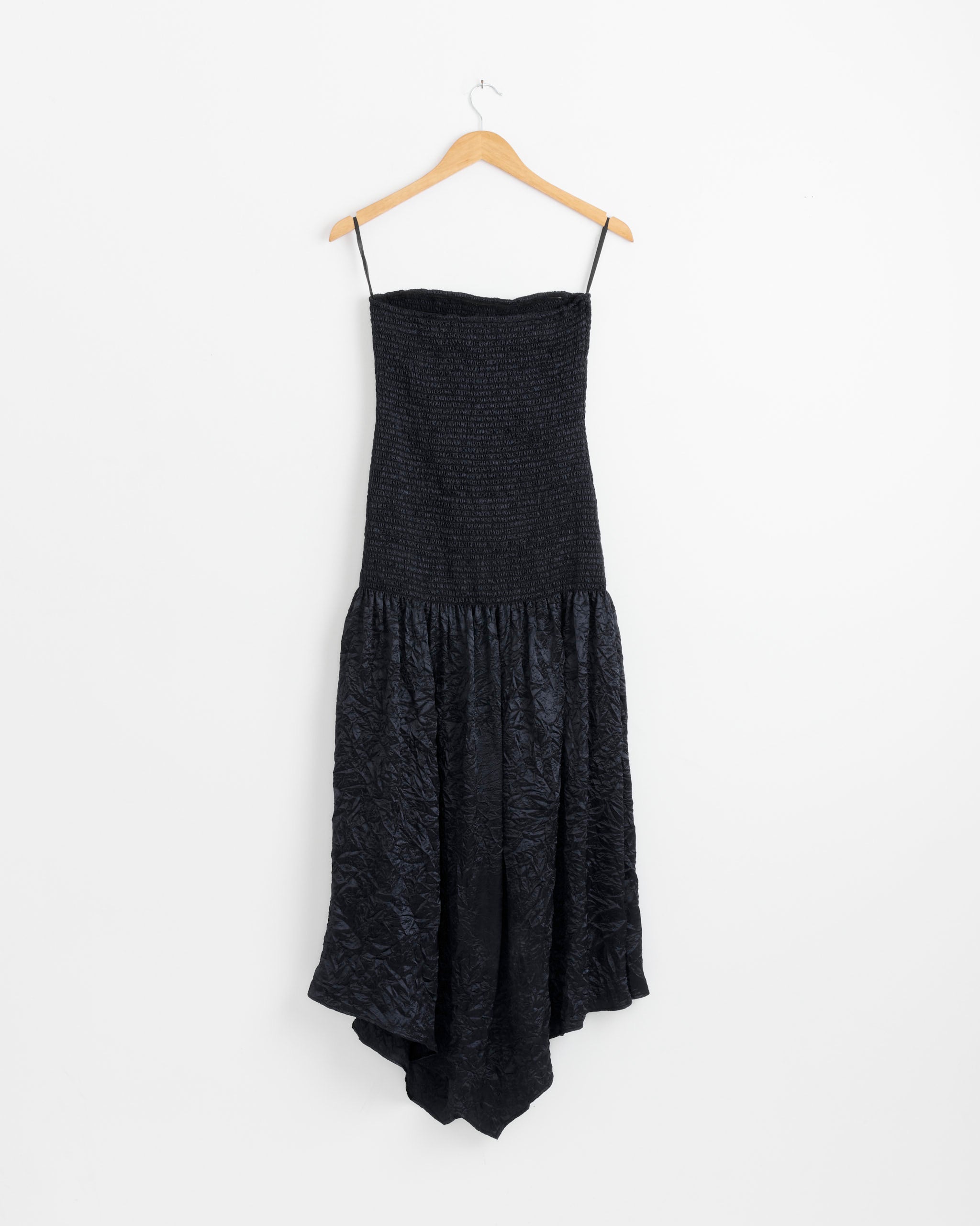 The NOMIA Smocked Strapless Maxi Dress in Midnight, featuring thin straps, a fitted smocked bodice, and a flared asymmetrical crinkle satin skirt, hangs on a wooden hanger against a plain white background.