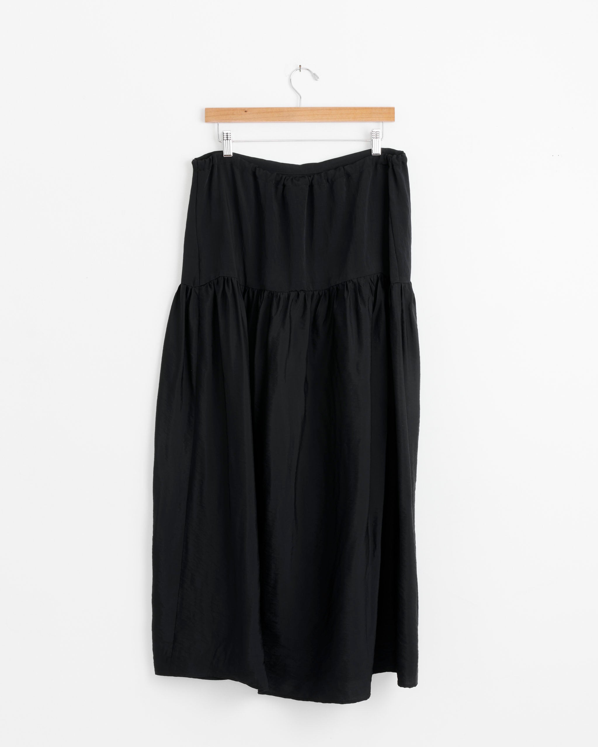 The NOMIA Maxi Gathered Skirt With Slit in Black, featuring a high waist and gathered waistband, hangs on a wooden hanger against a plain white background.