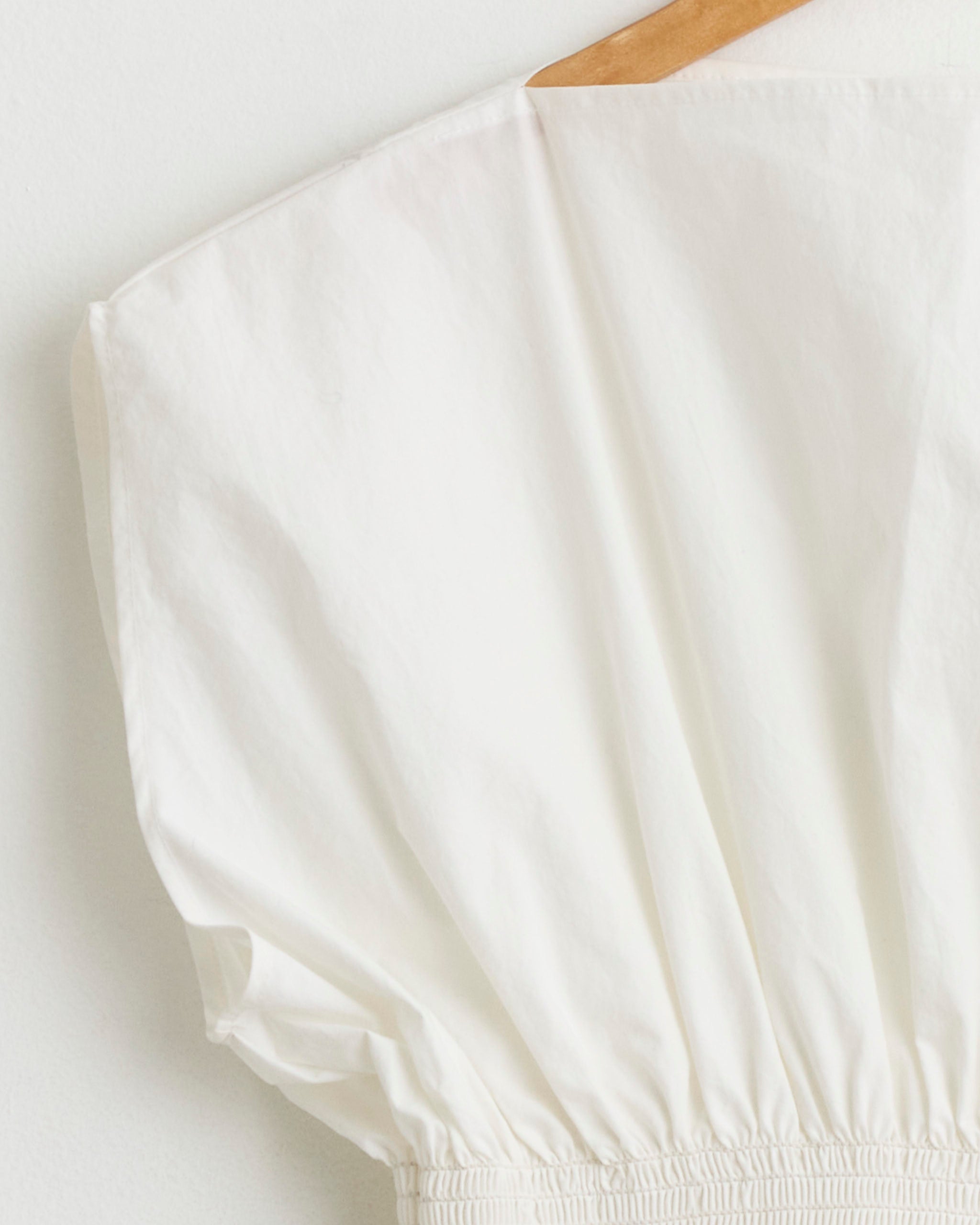 The NOMIA Smocked Sleeveless Top in Chalk, made in New York from washed cotton, features puffed sleeves, elastic-gathered hem, and hangs on a wooden hanger against a light background.