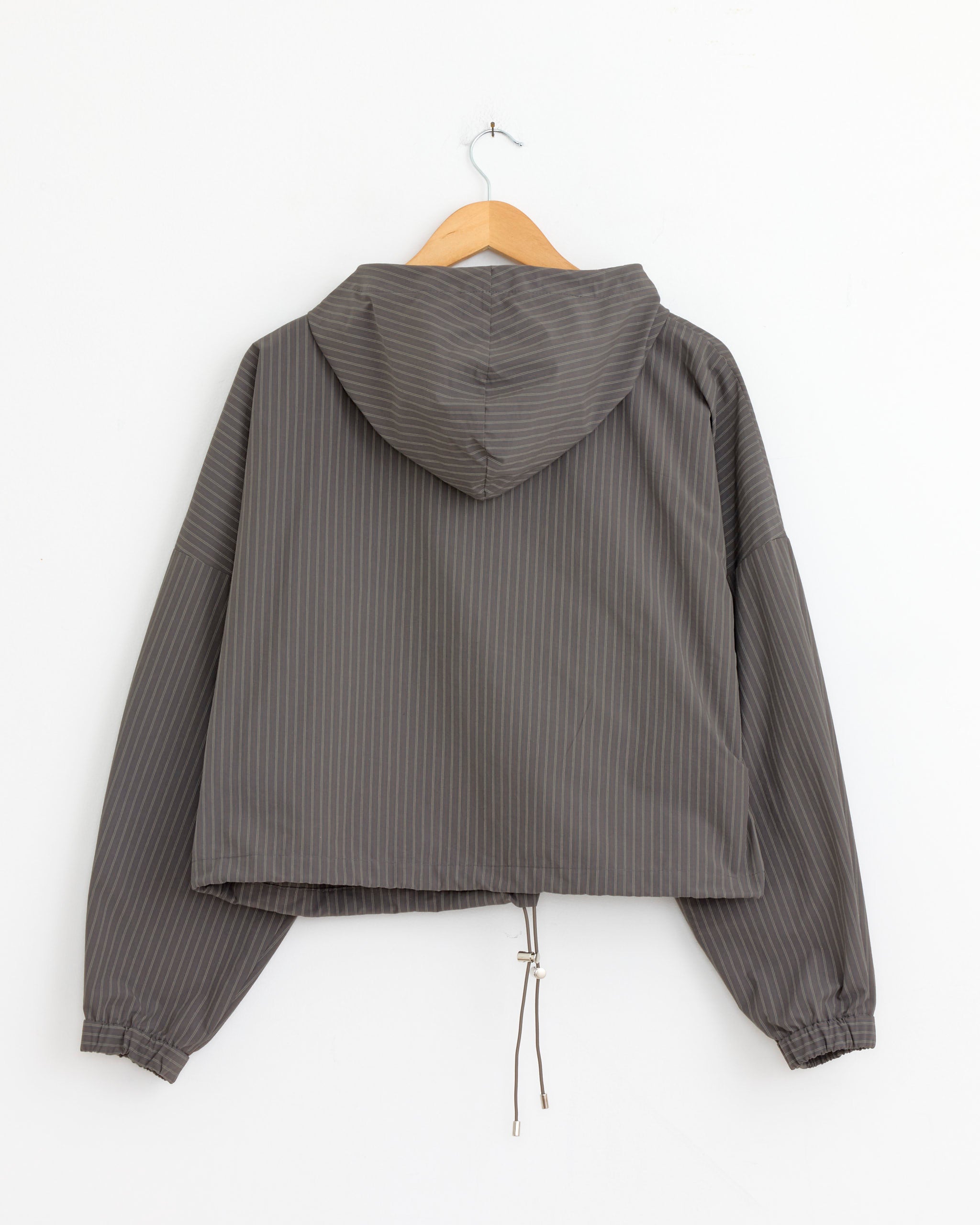 The NOMIA Zip Hooded Pullover in Coffee Stripe features gray vertical pinstripes, long sleeves, a zip front, adjustable drawstring hem, and a relaxed cropped fit. It’s shown hanging on a wooden hanger against a white wall.