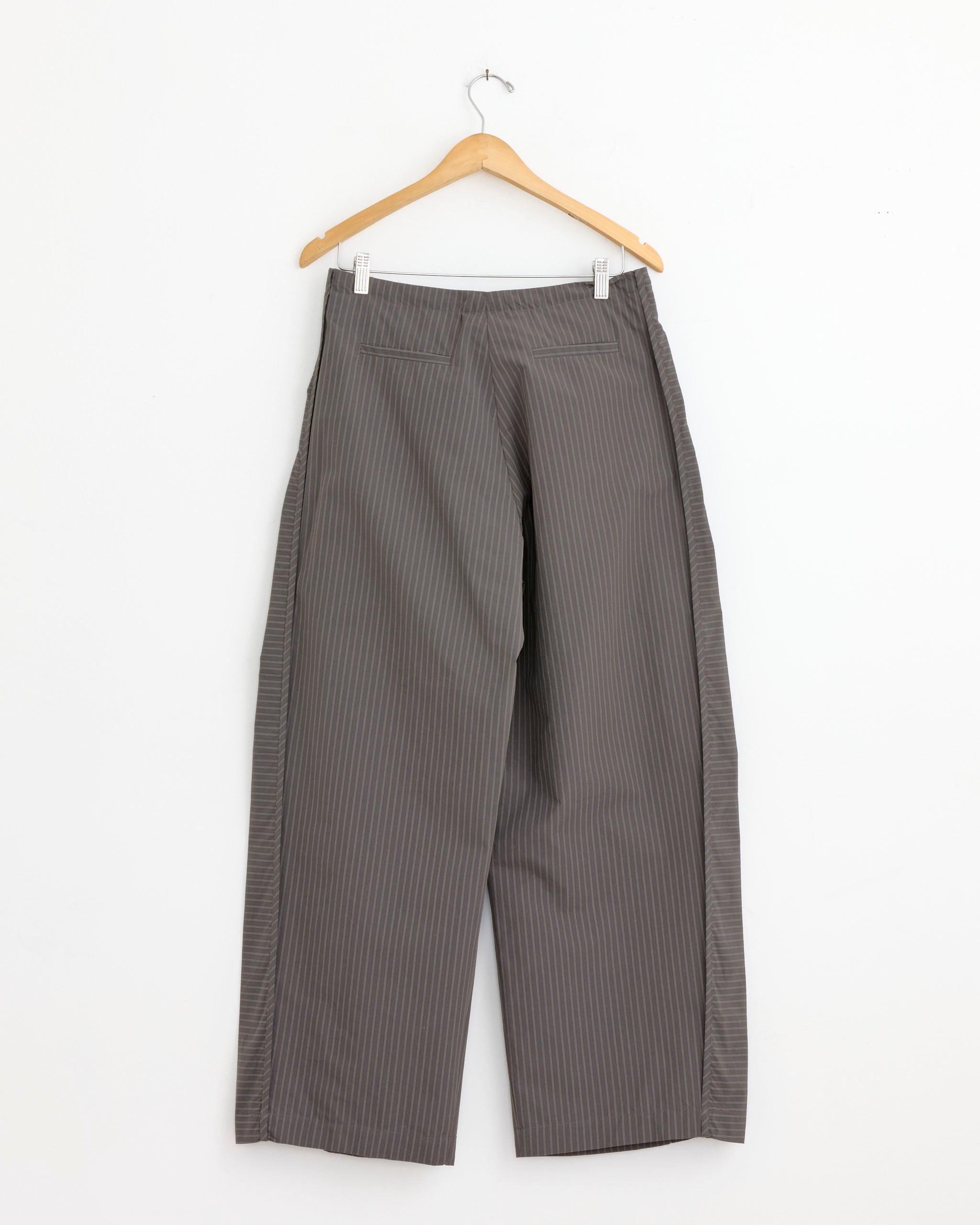 The NOMIA Side Panel Trouser in Coffee Stripe, wide leg with a drawcord waist, hangs on a wooden hanger against a white wall.