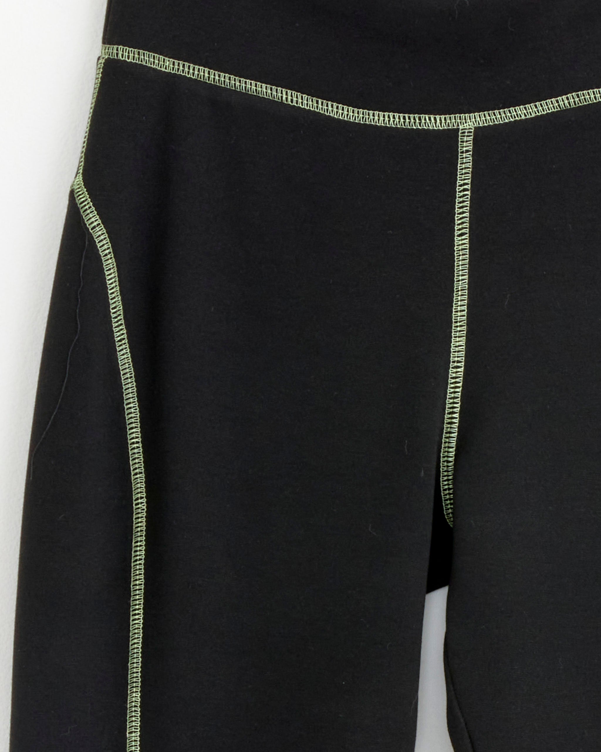 Close-up of NOMIA’s Scuba Capri Pant in Pistachio, black athletic leggings with green contrast stitching along the seams and waistband, displayed against a plain white background.