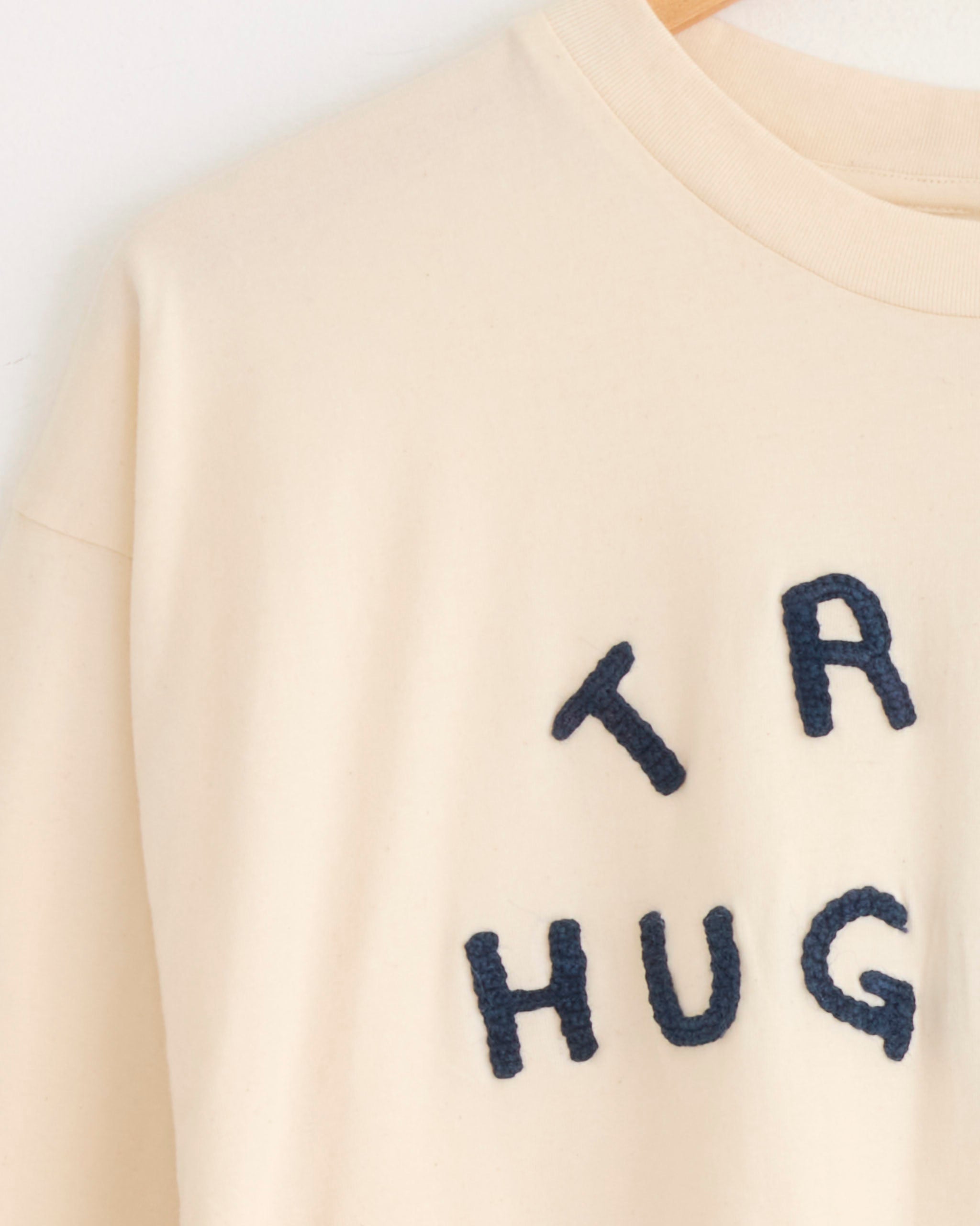 A close-up of the Story mfg Grateful Tee in Ecru Tree Hugger, hanging on a wooden hanger with dark blue textured “TR HUG” lettering in the signature style.