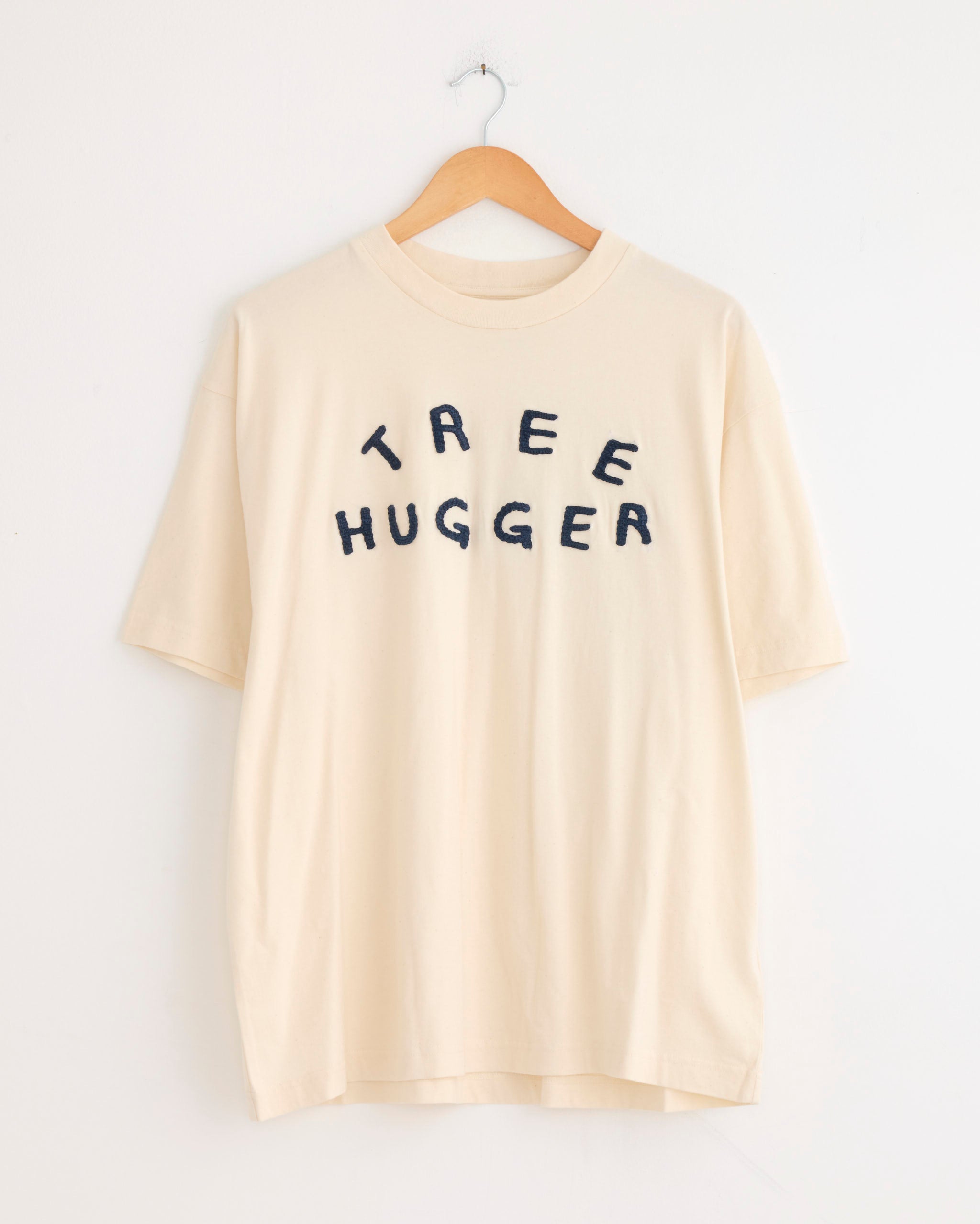 The Story mfg Grateful Tee in Ecru Tree Hugger, crafted from organic cotton, hangs on a wooden hanger and features bold, uneven black "TREE HUGGER" lettering across the chest.