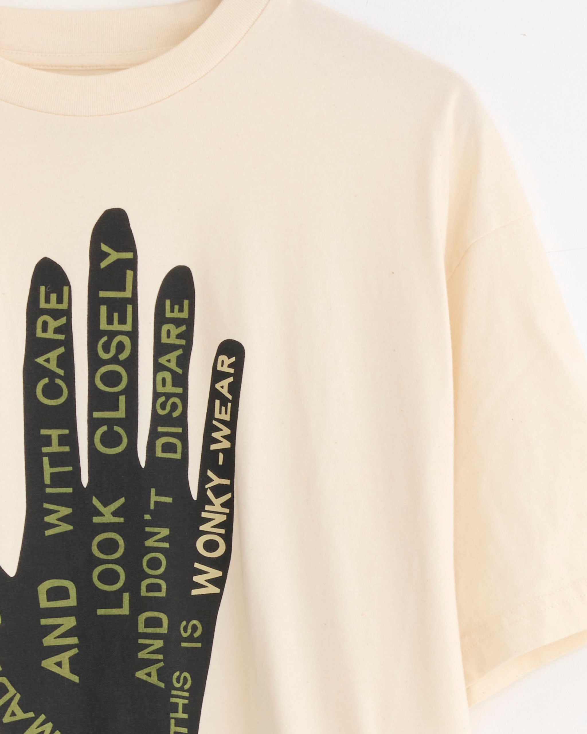 The Story mfg Grateful Tee in Made Slowly is an organic cotton cream tee with a bold black hand graphic and green/black text on the front reading "HANDLE WITH CARE AND LOOK CLOSELY AND DONT DESPAIR THIS IS WONKY-WEAR." Vintage-inspired fit.
