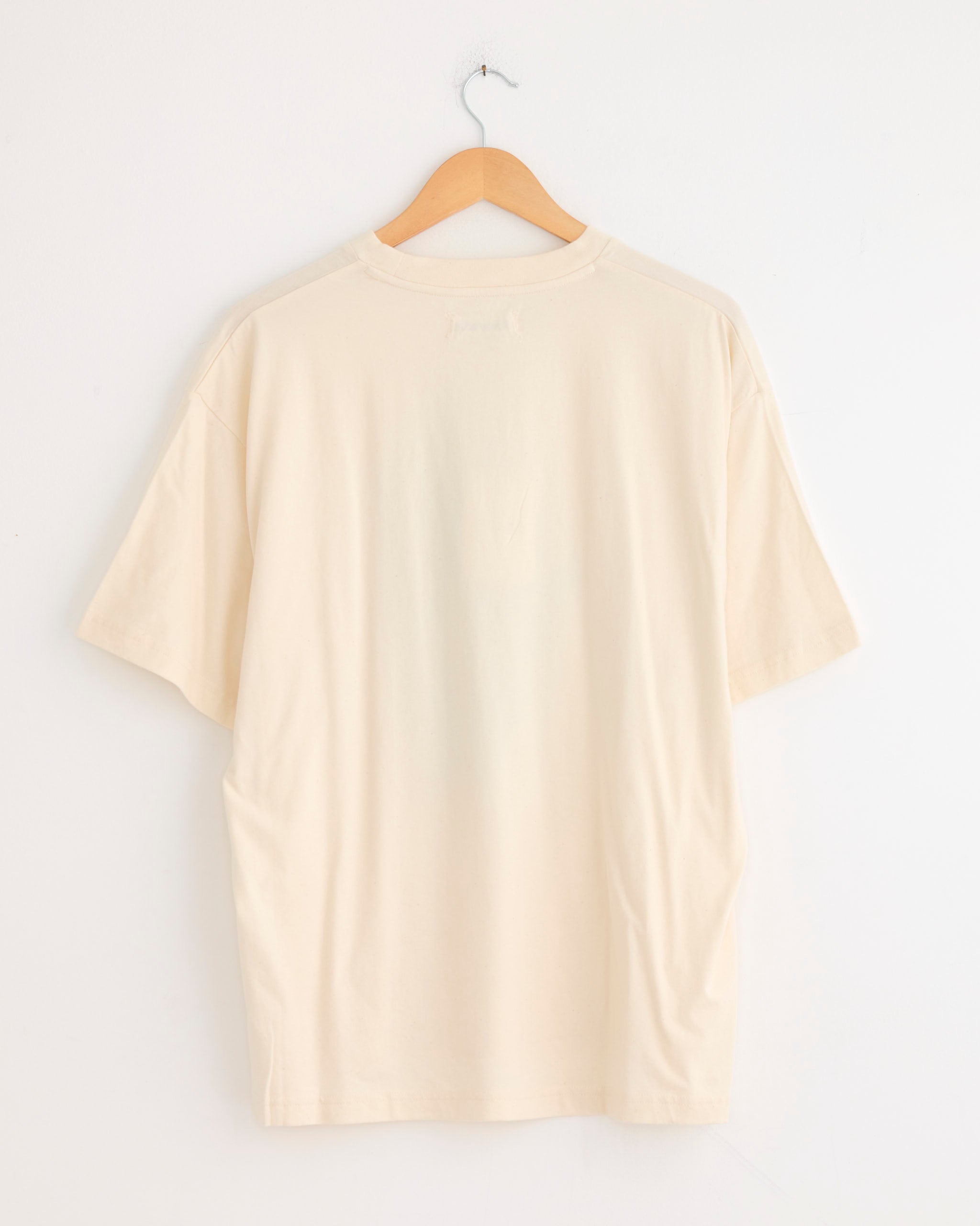 The Grateful Tee in Made Slowly by Story mfg is a short-sleeved, off-white organic cotton T-shirt with a subtle vintage-inspired fit, shown from the back on a wooden hanger against a white wall.