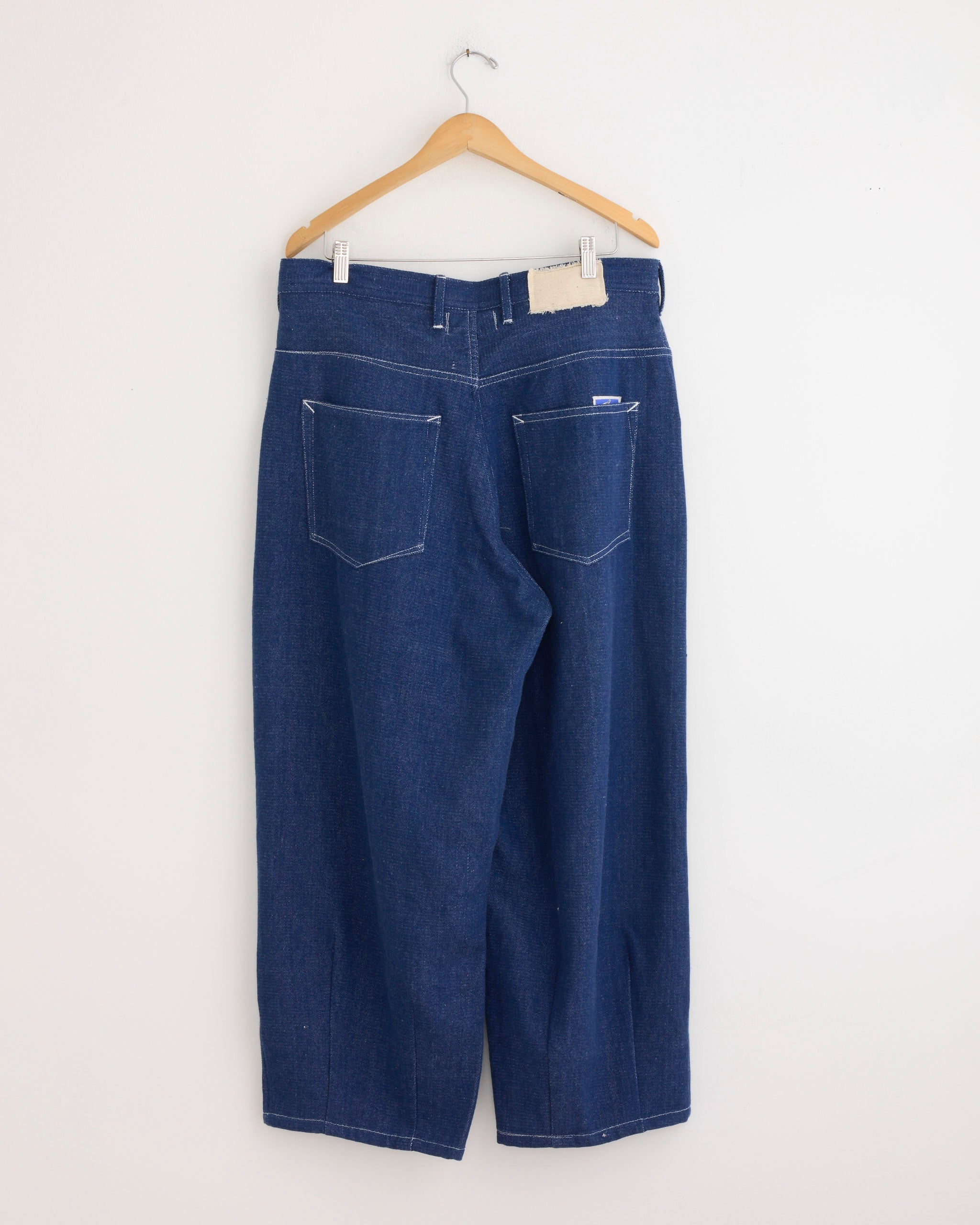 The Lush Jean in Handloom Denim by Story mfg hangs on a wooden hanger against a plain white wall, showcasing its wide-leg cut, white stitching, and two large back pockets.