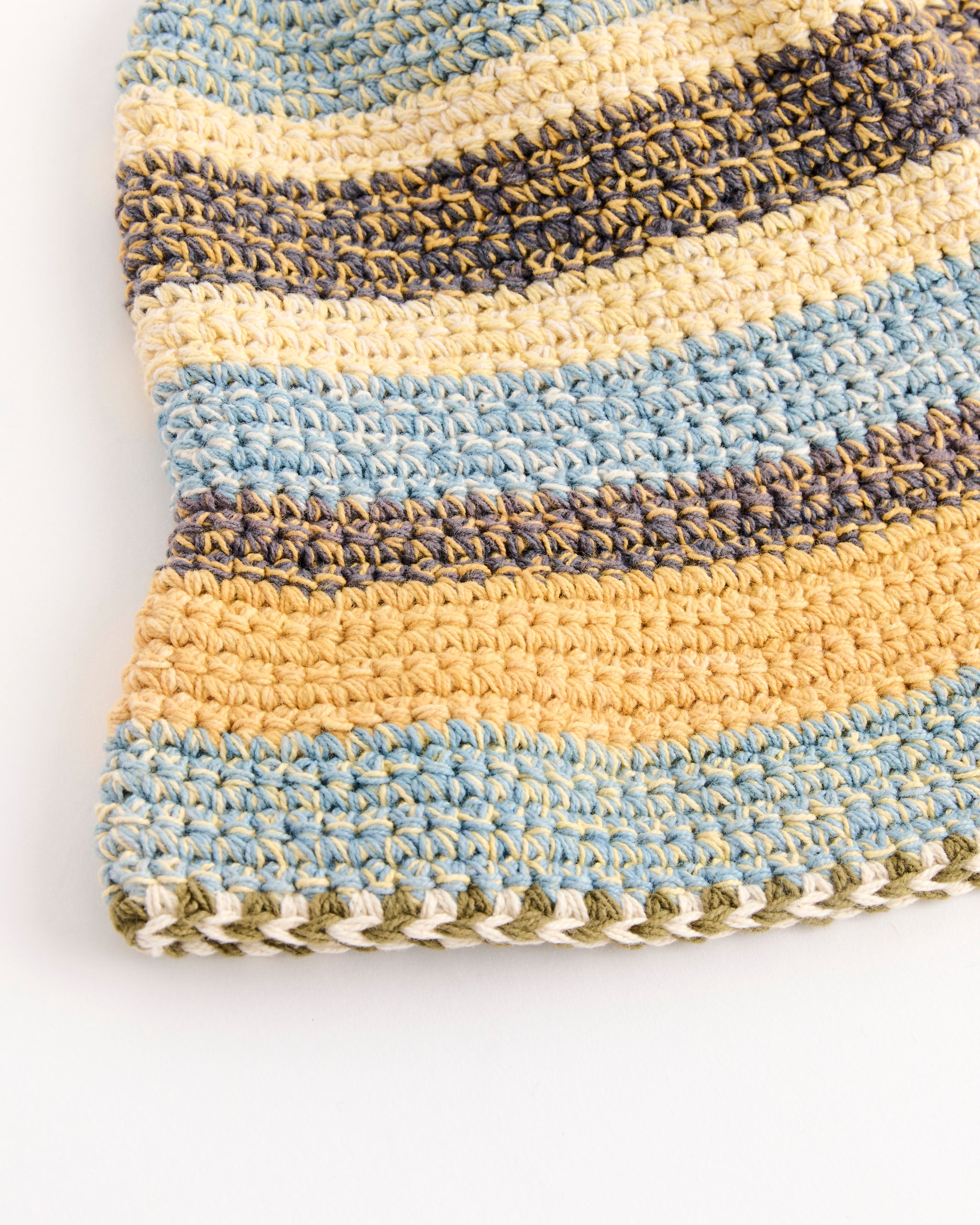 A close-up of the Story mfg Brew Hat in Twisted Stripe, hand-crocheted from organic cotton with horizontal blue, yellow, beige, and gray stripes on white. Naturally dyed fibers and stitch details are clearly visible.