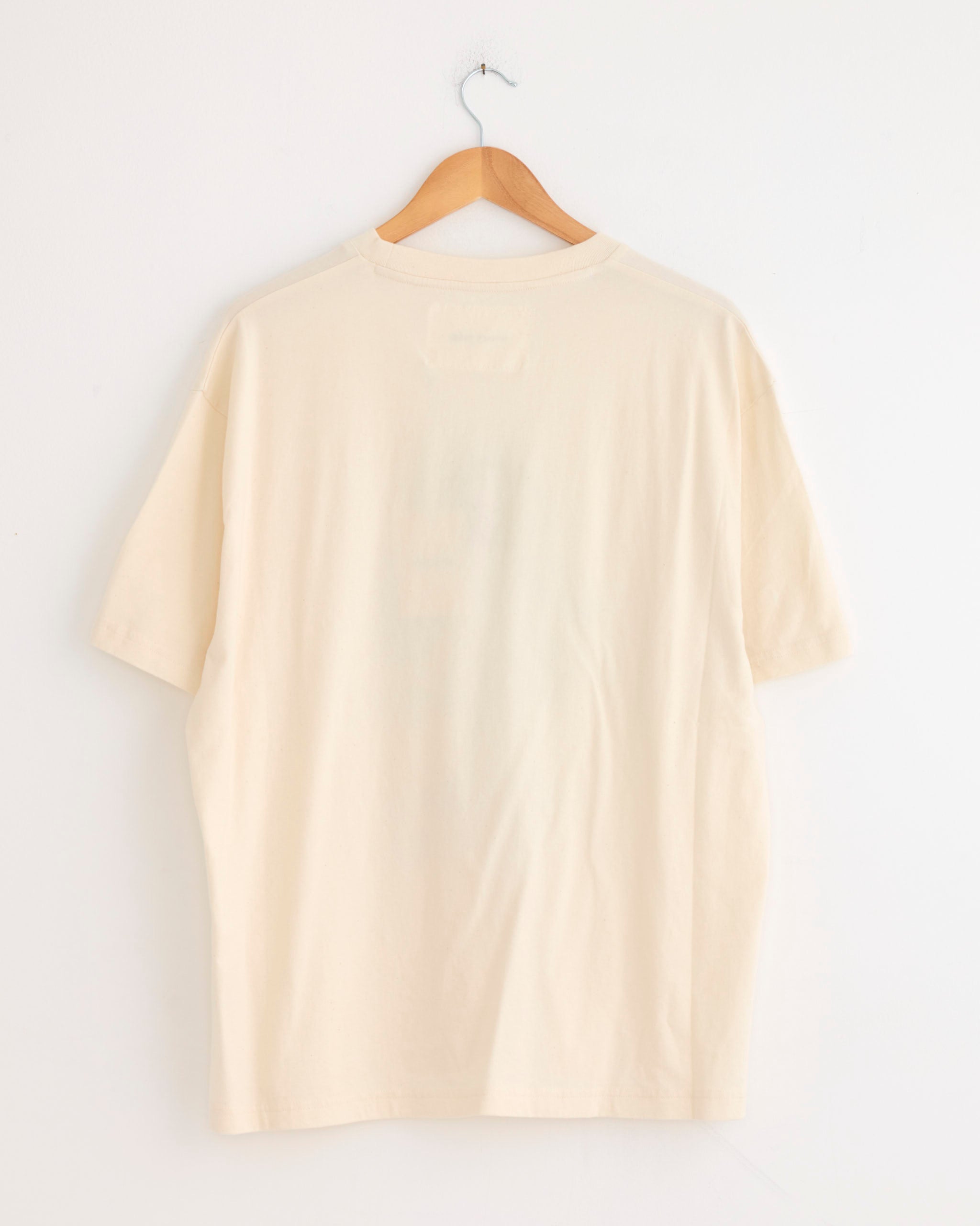 The Story mfg Grateful Tee in Young Trees, a plain off-white short-sleeve shirt made from organic cotton, hangs on a wooden hanger against a white wall, displayed from the back to highlight its vintage-inspired fit.