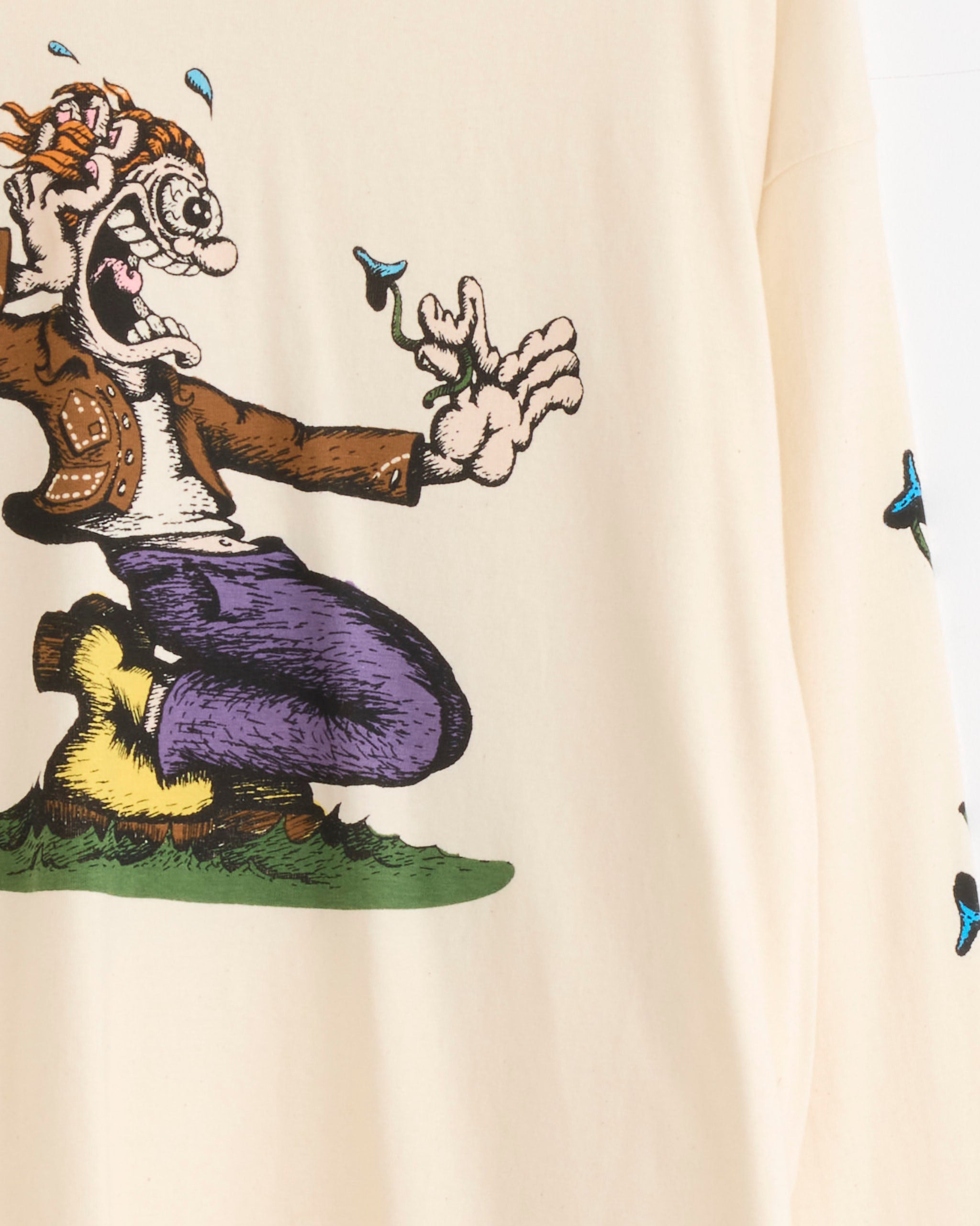 A wide-eyed character kneels on grass wearing the Story mfg Grateful Tee in The Trip, with blue flowers sprouting from their fingers as they reach out, dressed in a vintage-inspired fit.
