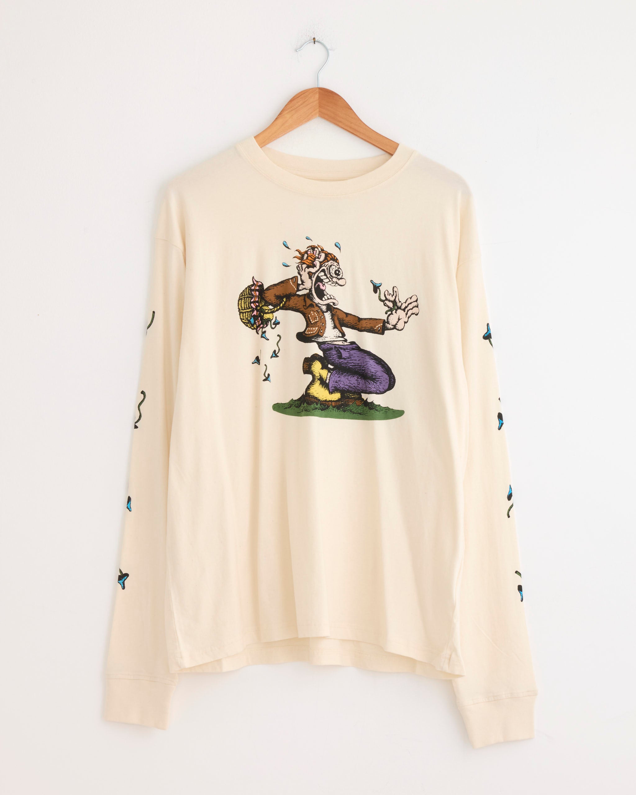 The Grateful Tee in The Trip by Story mfg is a cream-colored, organic cotton long-sleeve shirt with vintage-inspired sleeve details and a vibrant cartoon character kneeling on grass with music notes, displayed on a wooden hanger.