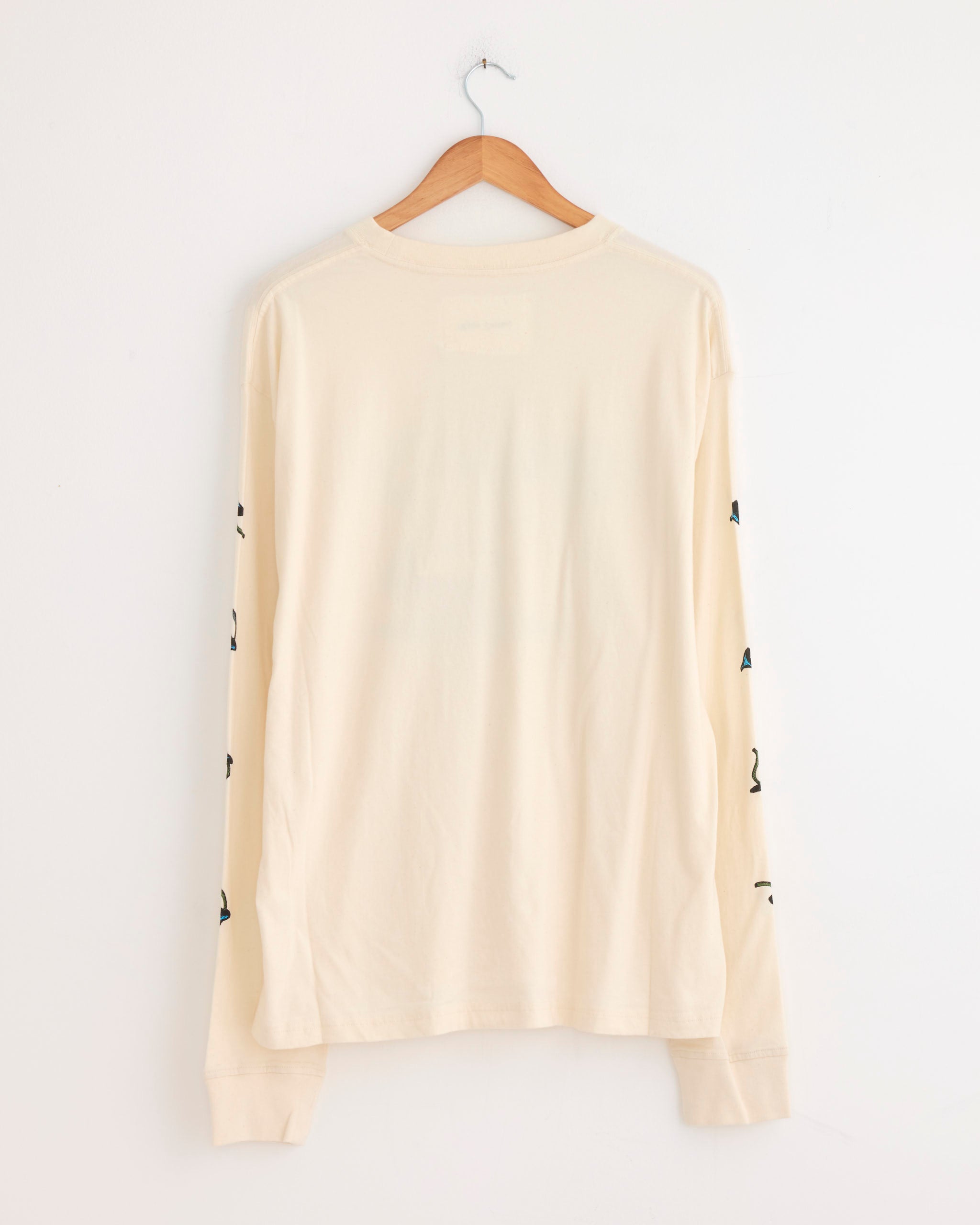 The Grateful Tee in The Trip by Story mfg is a long-sleeved, off-white organic cotton shirt with black graphics on the sleeves, shown from the back on a hanger. It features a vintage-inspired fit.