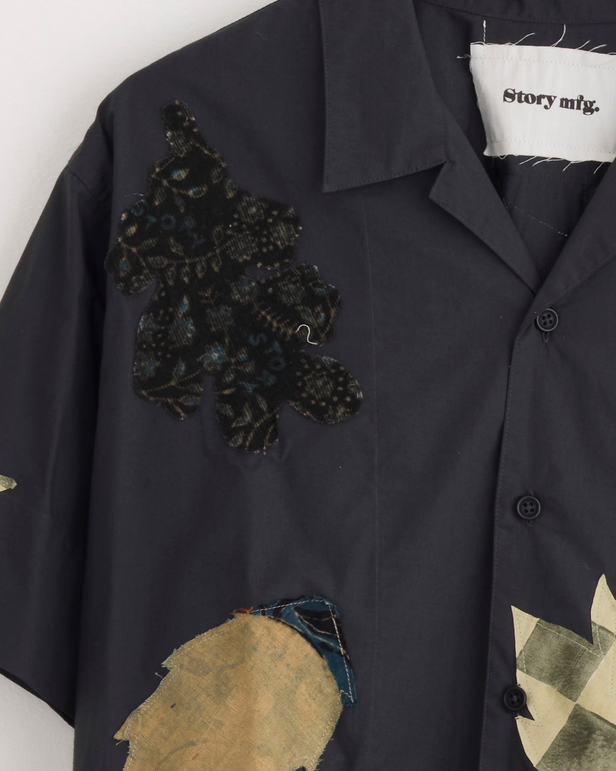 A close-up of the Story mfg Greeting Shirt in Charcoal Leaf Scatter, an organic cotton shirt with sewn fabric patches: a black patterned patch on the shoulder and beige shapes on the chest. The inside label reads Story mfg.