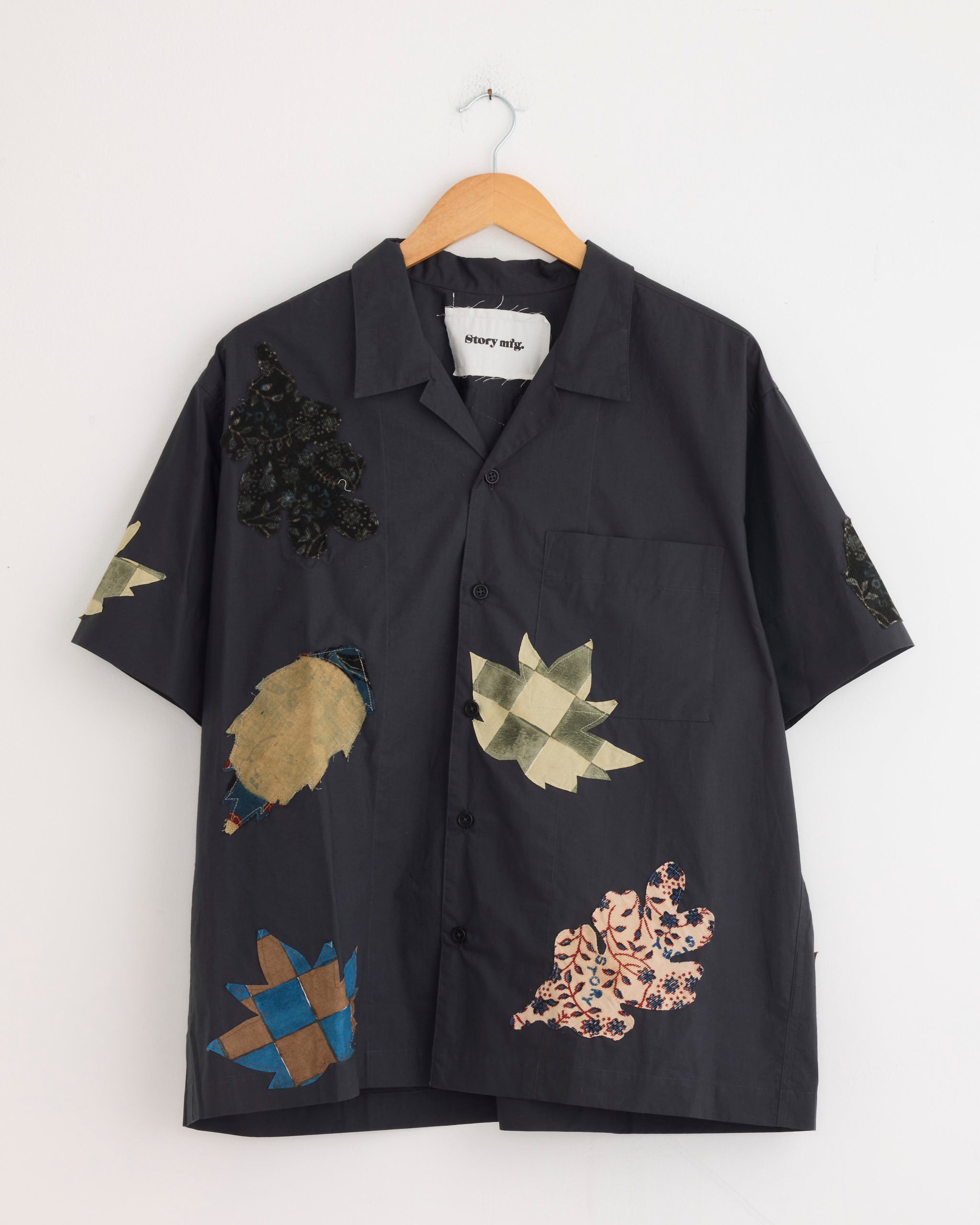 The Story mfg Greeting Shirt in Charcoal Leaf Scatter is an organic cotton, short-sleeve button-up with a front pocket and colorful leaf patches on the front and sleeves, shown on a wooden hanger.