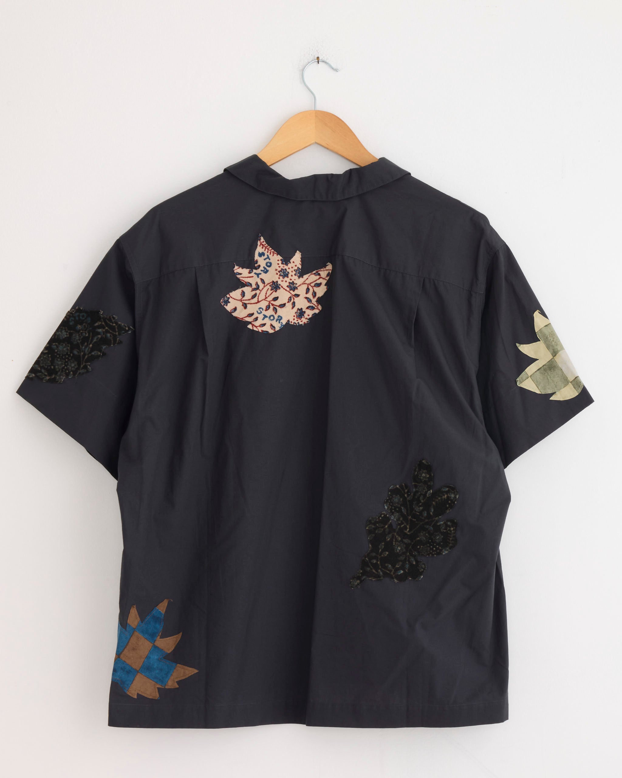 The Story mfg Greeting Shirt in Charcoal Leaf Scatter, made from organic cotton, hangs on a hanger against a white wall. It features colorful appliqué quilt patterns in assorted shapes and fabrics on the back.