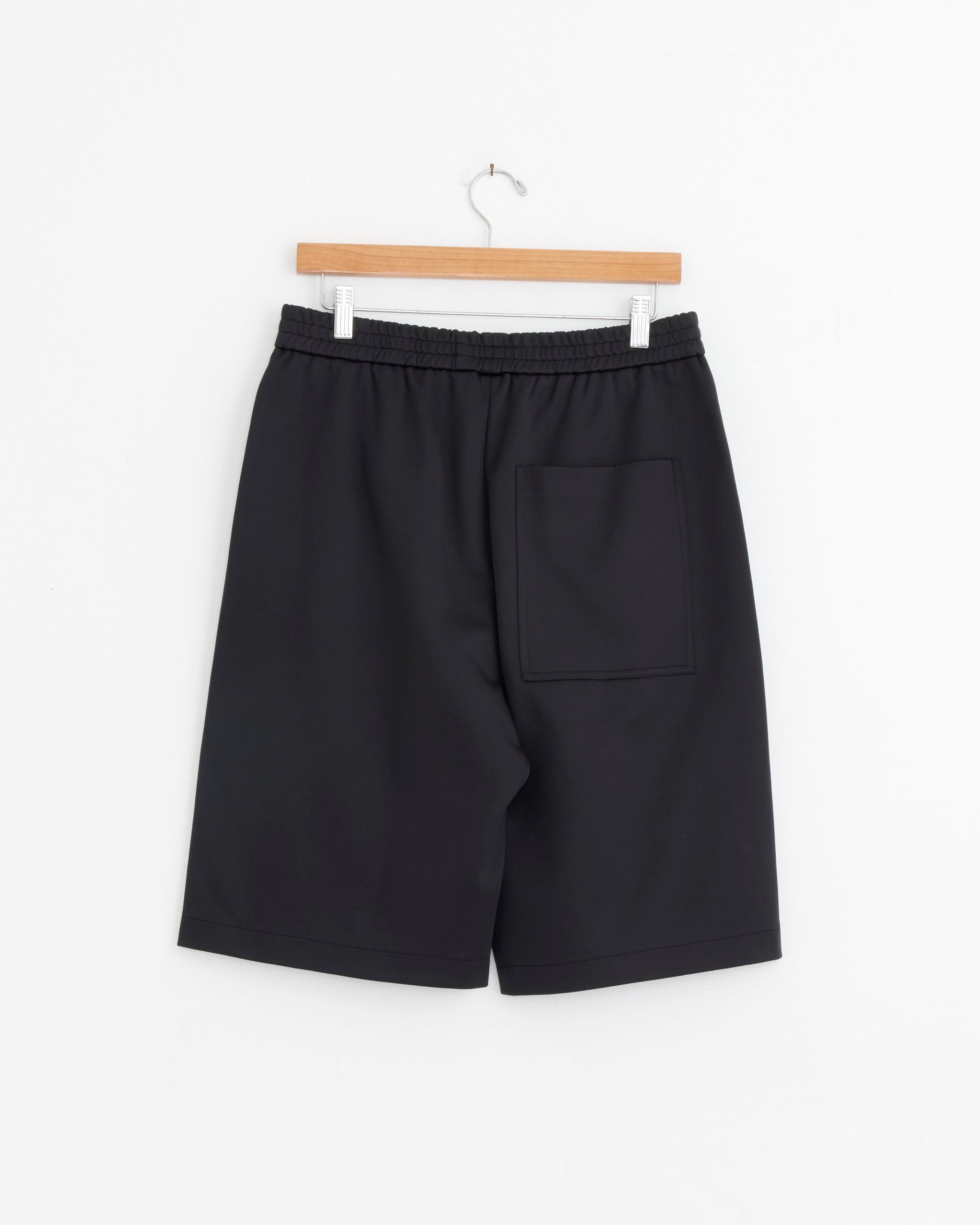 The Tibi Active Knit Short in Black, crafted from soft Italian poly-cotton knit with an elastic waistband and a back patch pocket, is shown knee-length on a wooden hanger against a plain white background.