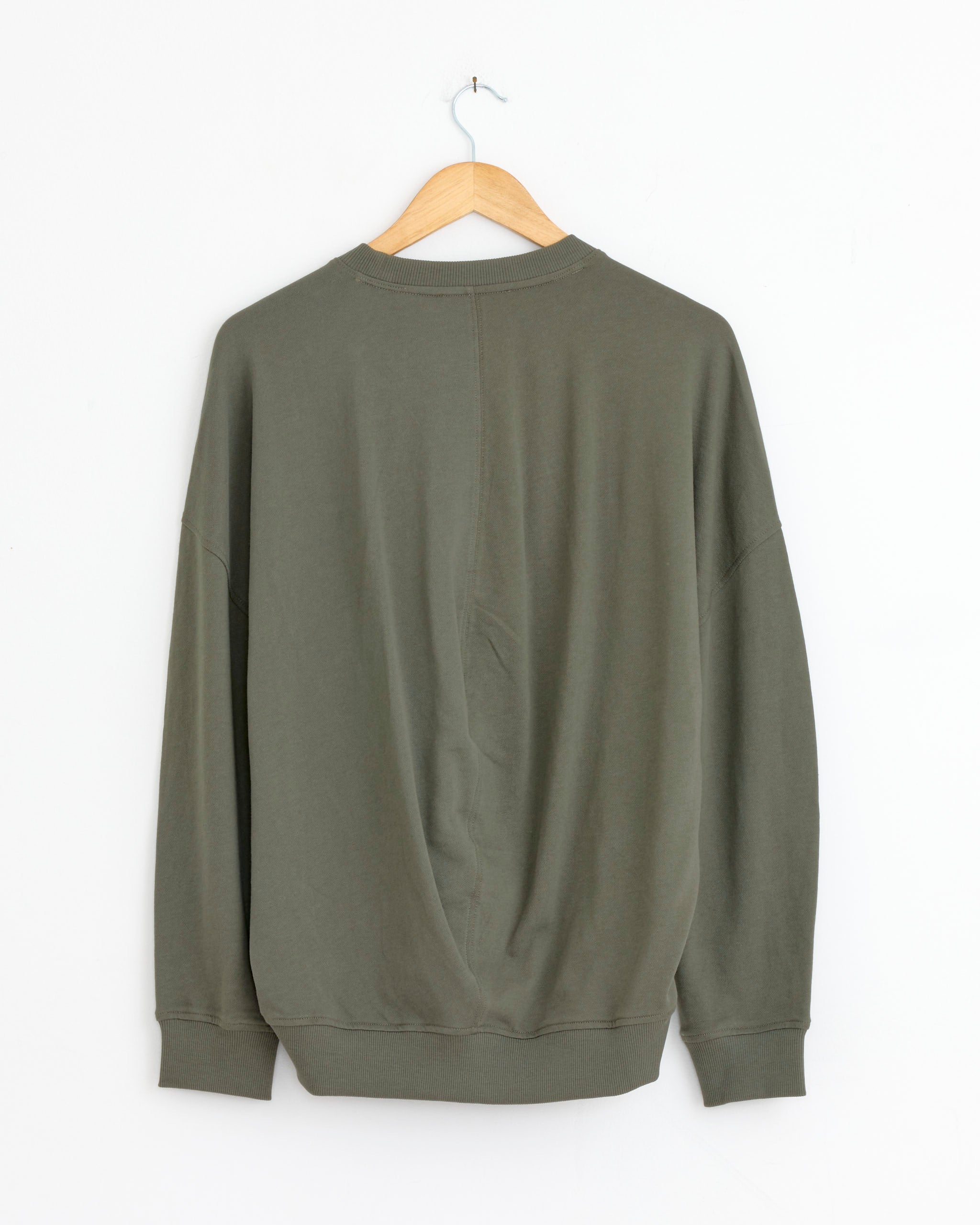 The Tibi Light Weight Sweatshirt in Olive, made from soft Peruvian cotton for comfort, is displayed on a wooden hanger against a white wall with its back facing outward.