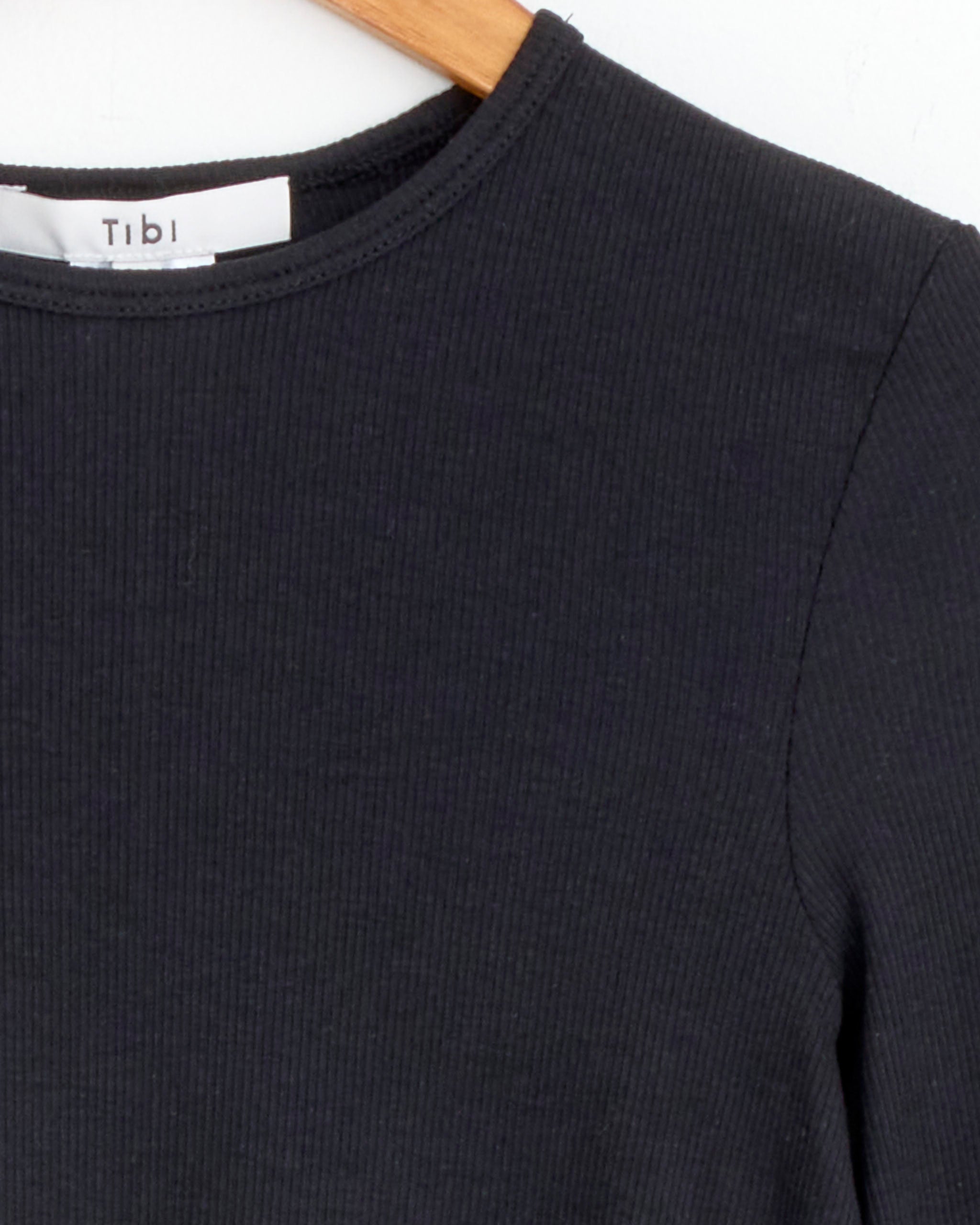 A close-up of the Tibi Circular Ribbed Pullover in Black, made from a Peruvian cotton blend, hangs on a wooden hanger, highlighting its crewneck and shoulder details with a visible partial Tibi label.