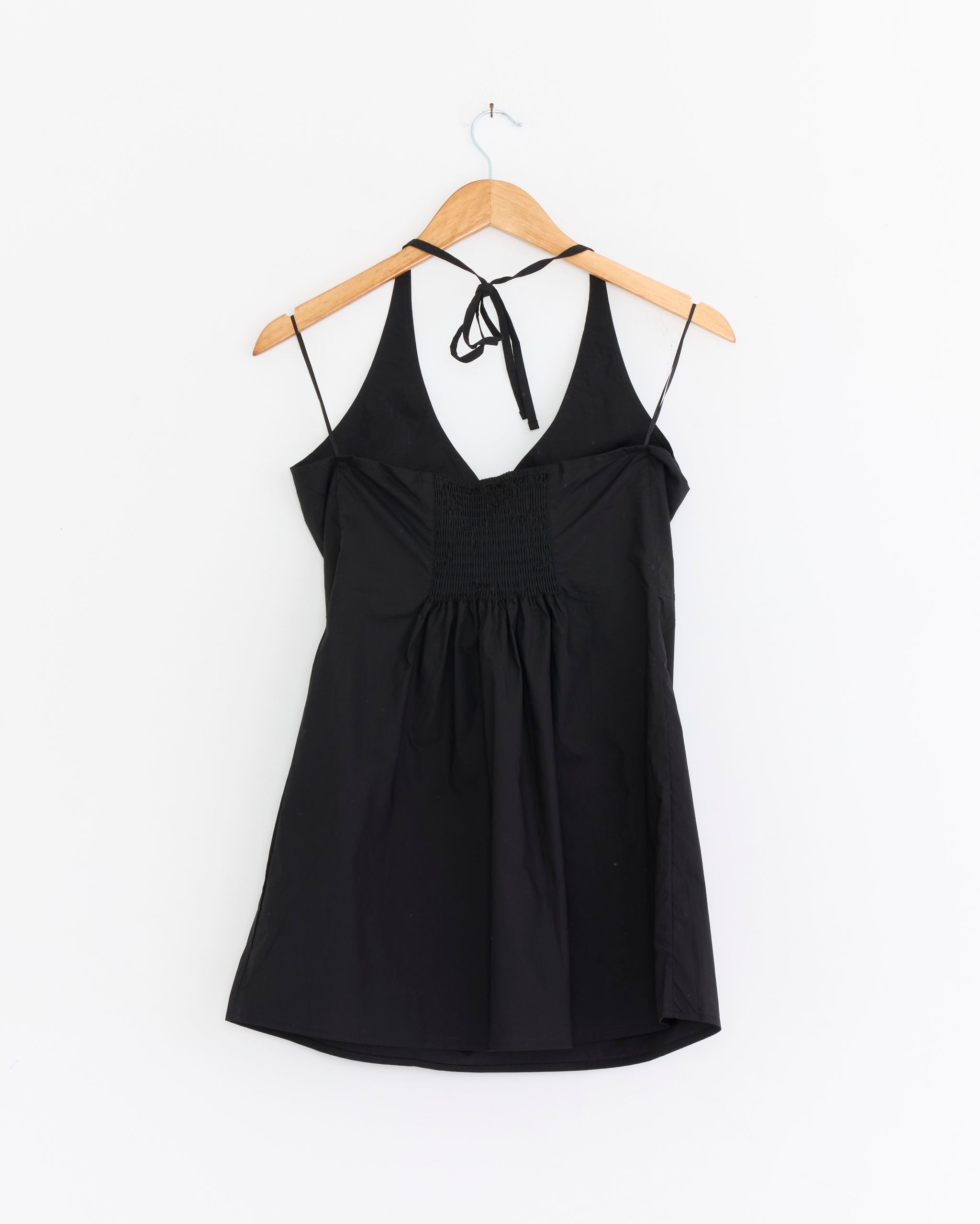 The Deiji Studios Cinched Halter Dress in Black, featuring a gathered front detail, hangs on a wooden hanger against a plain white wall, embodying the brand’s signature effortless style.
