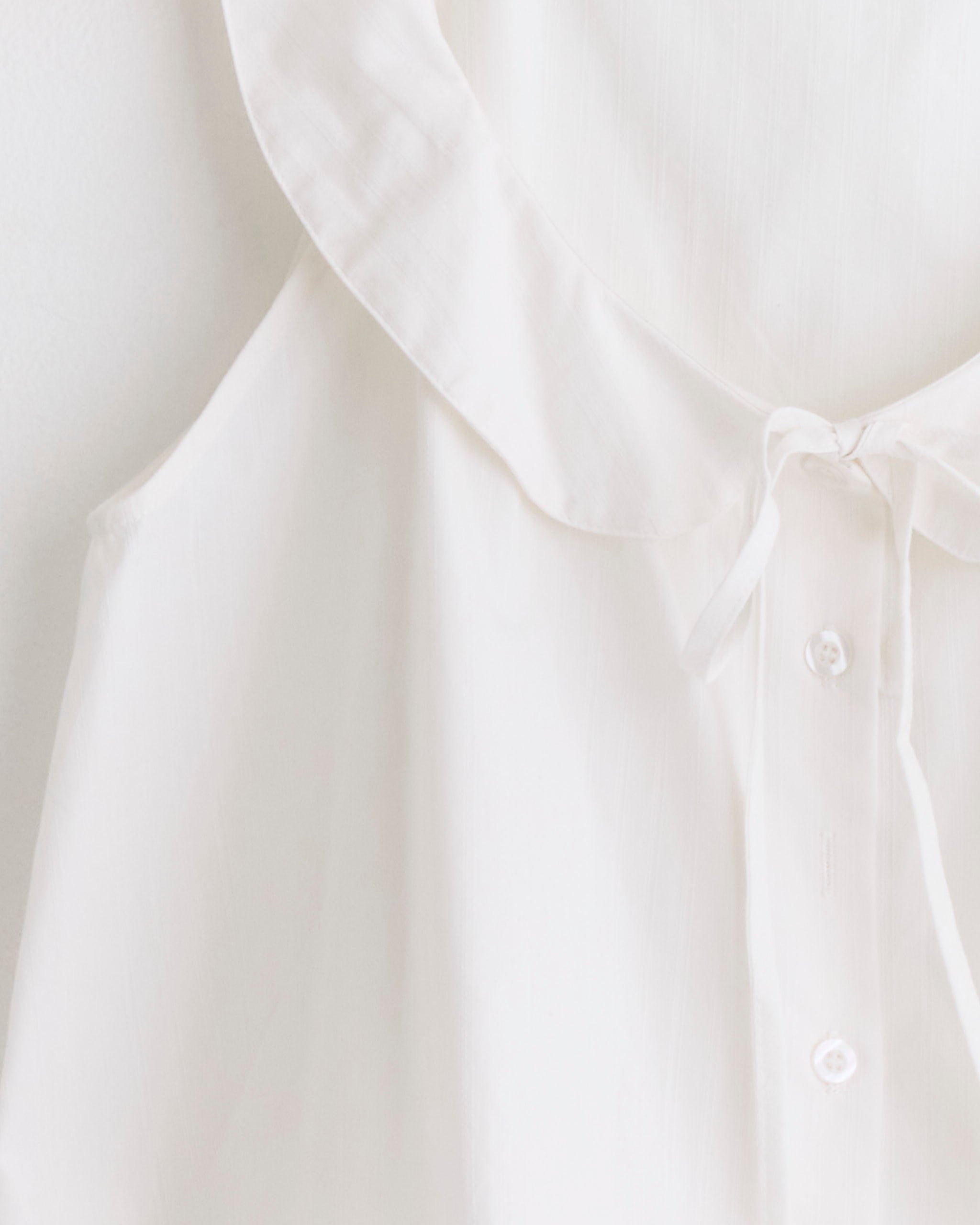 A close-up of the Deiji Studios Round Collar Set in White Texture, a sleeveless blouse with a ruffled collar, button-down front, and tie neckline, crafted from lightweight, slightly sheer organic cotton for an airy feel.