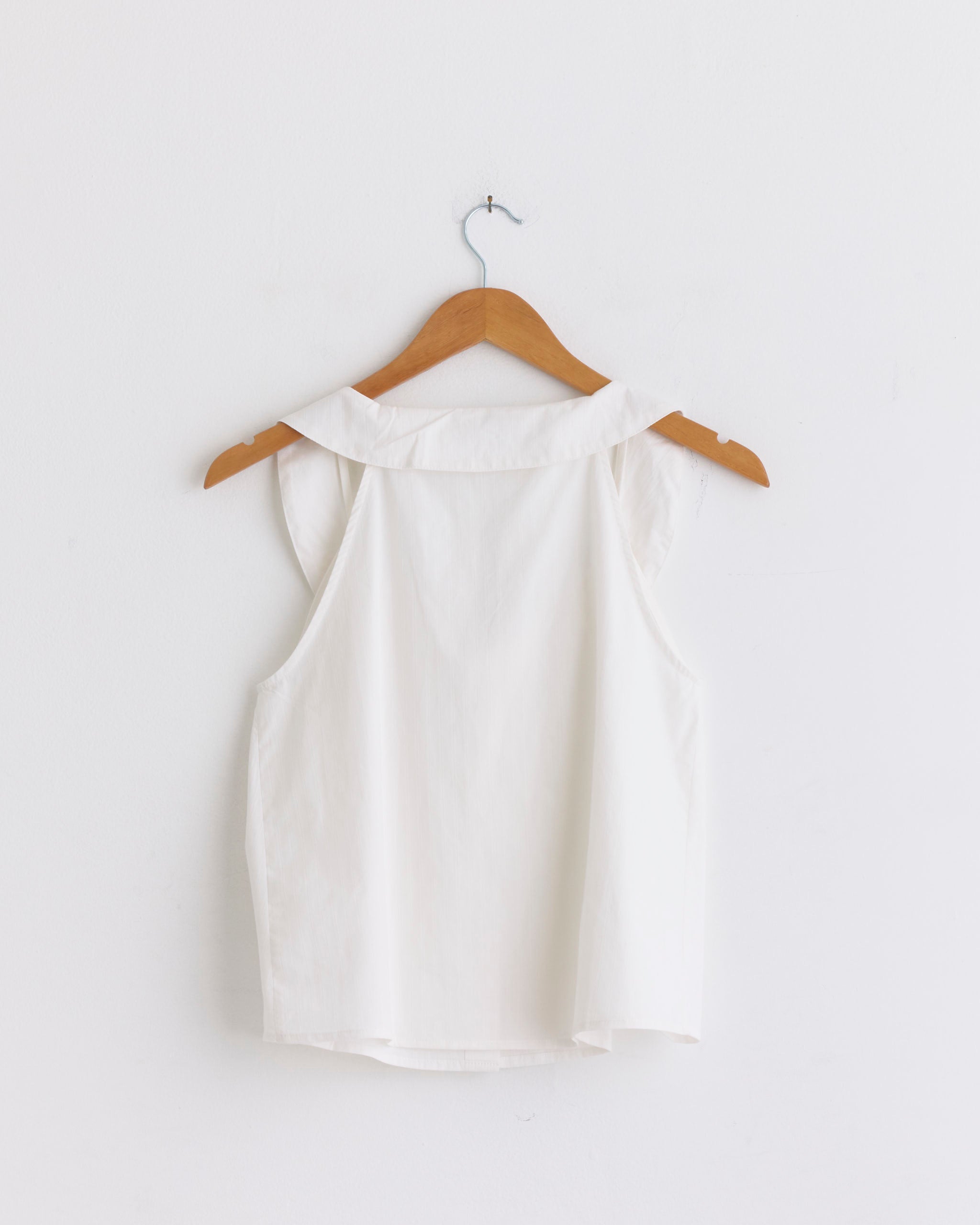 The Round Collar Set in White Texture by Deiji Studios features a sleeveless organic cotton blouse with a wide collar, displayed on a wooden hanger against a plain white wall—an elegant nod to the brand's effortless style.