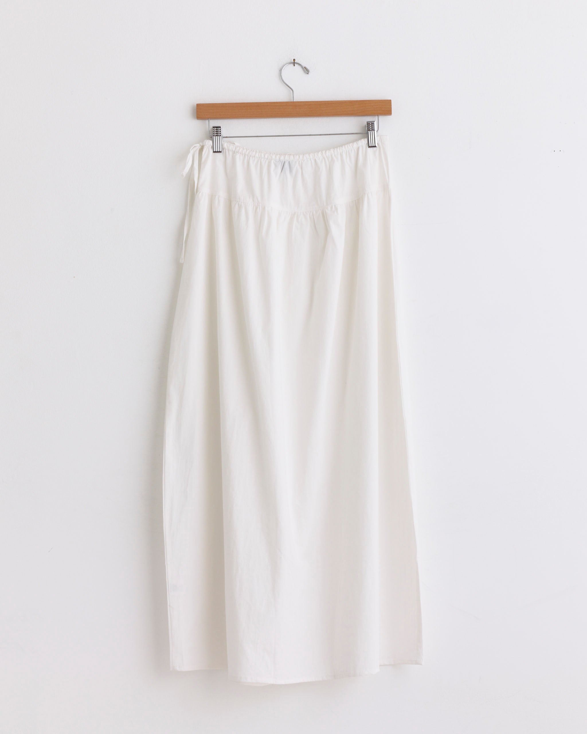 The Maxi Skirt in White Texture by Deiji Studios—a long, plain organic cotton skirt with an elastic waistband—hangs on a wooden hanger against a white wall.