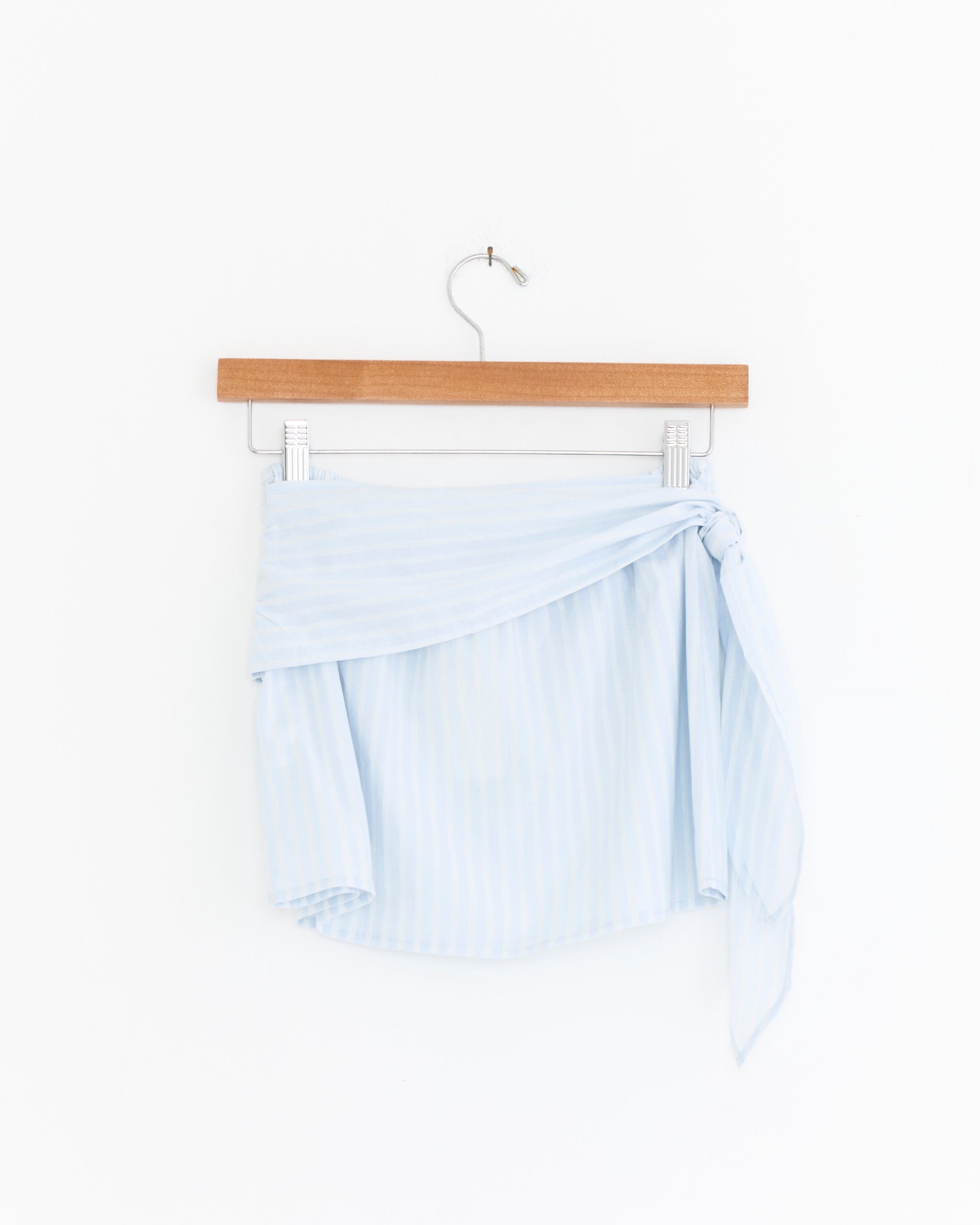 The Belted Skirt in Powder Blue Stripe by Deiji Studios, made from organic cotton with a side tie, is displayed on a wooden hanger against a plain white background.
