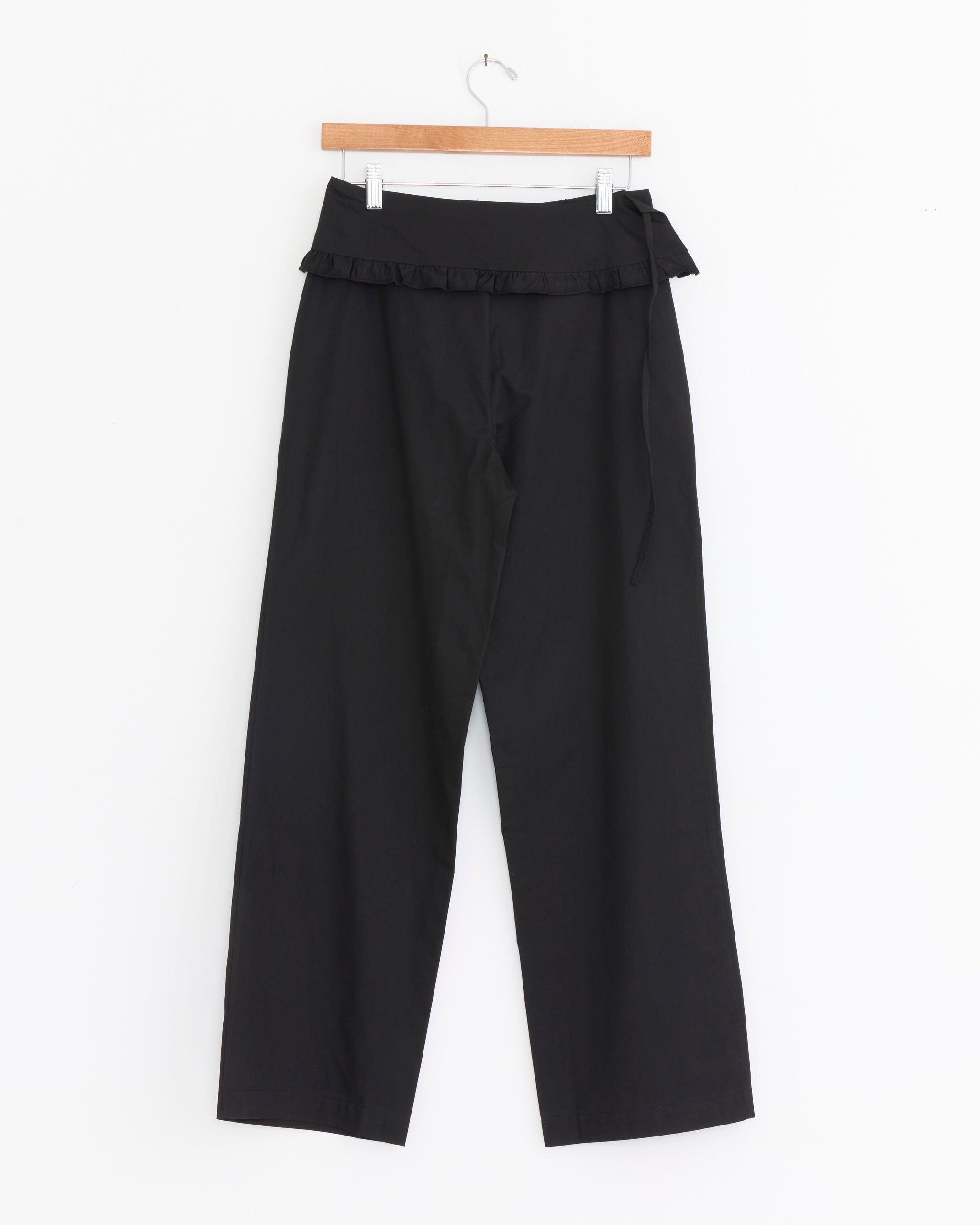 The Deiji Studios Flounce Belt Pant in Black features a high waist, wide-leg fit, ruffled trim, and side-tie detail, shown hanging on a wooden hanger against a white background.