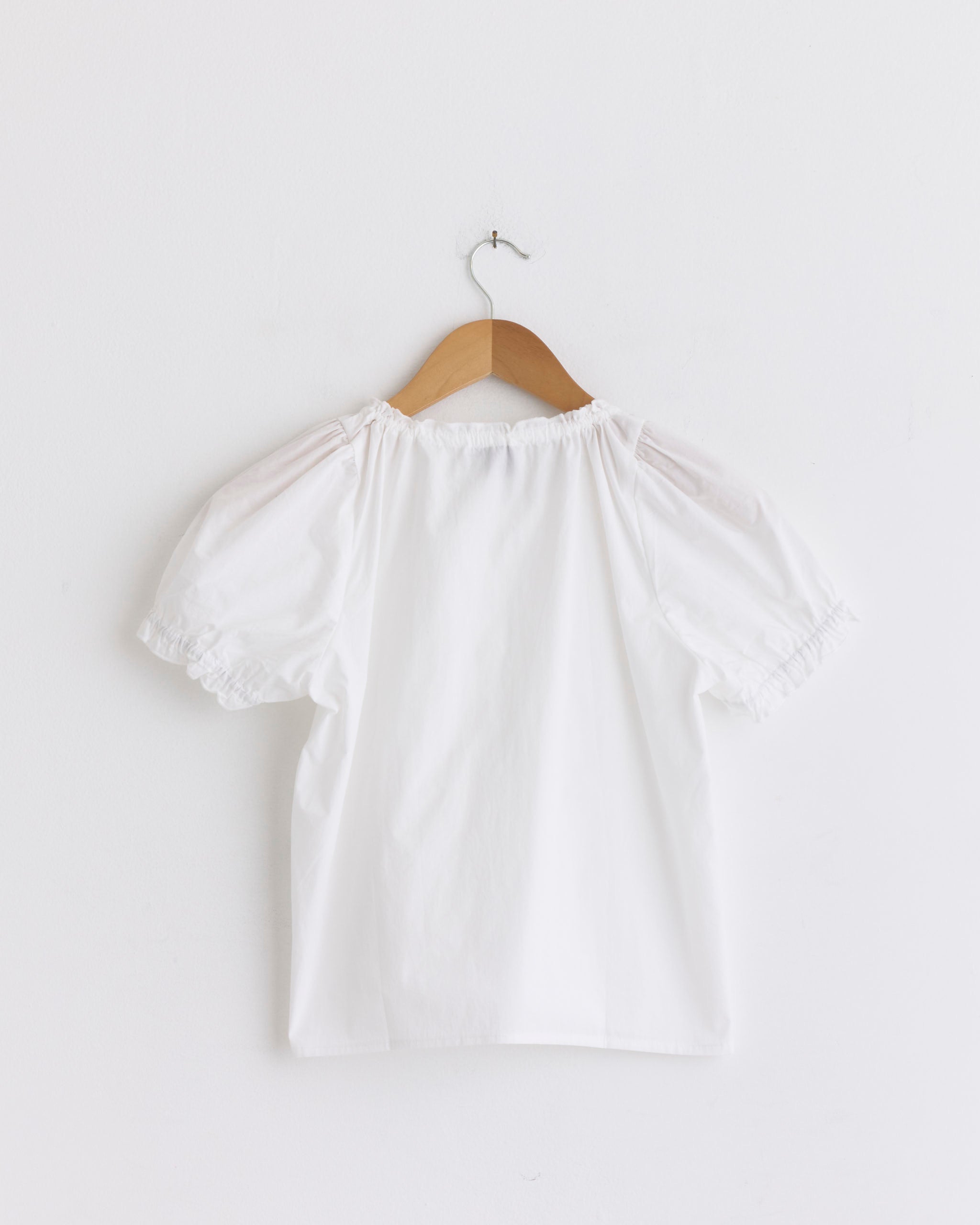 The Close Shirt in White by Deiji Studios, featuring short sleeves and ruffled cuffs, hangs on a wooden hanger against a plain white wall—delivering a fresh, airy update to the classic cropped relaxed shirt.