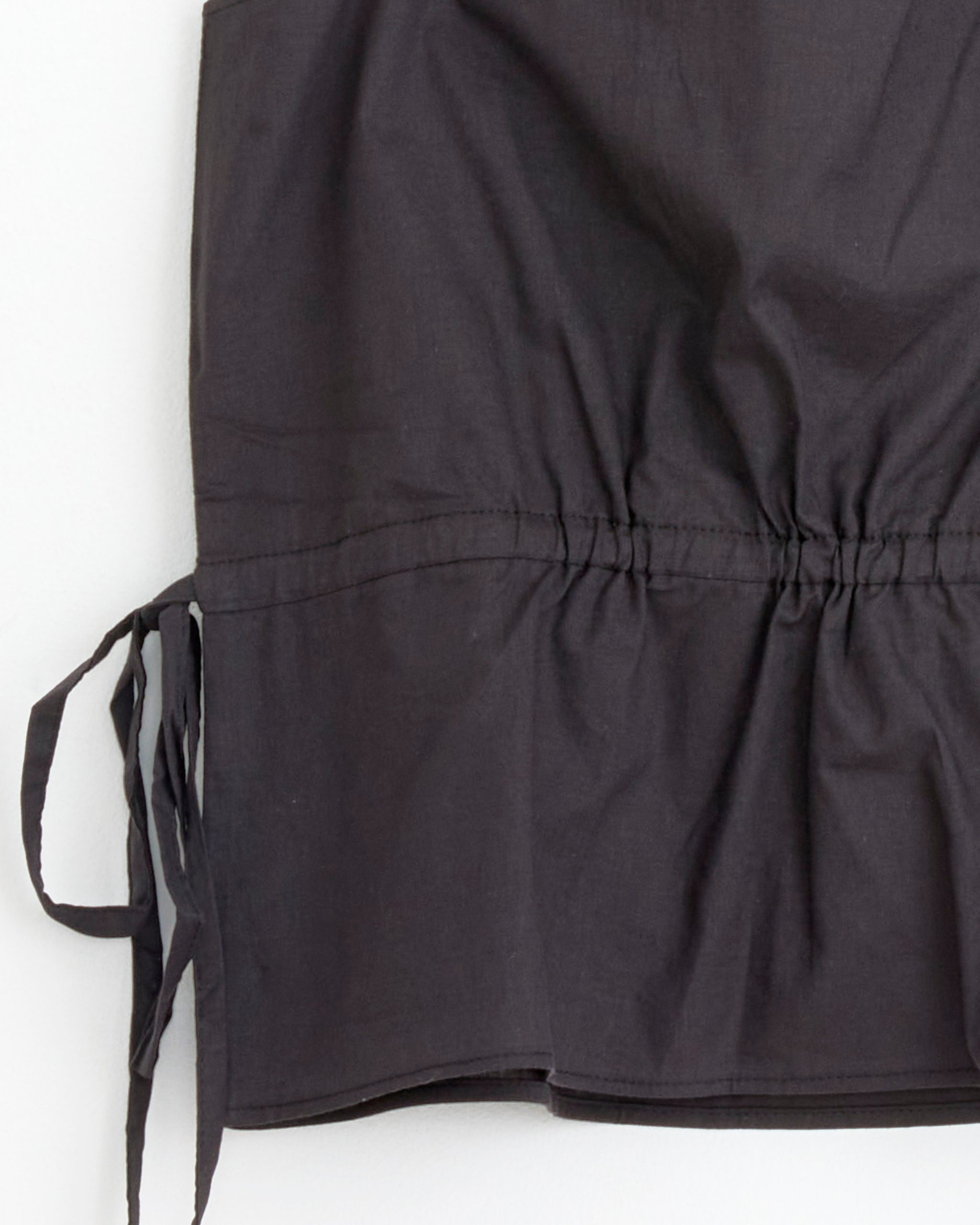 A close-up of Deiji Studios’ Waist Tie Top in Black, crafted from organic cotton voile with side ties and gathered stitching, shown against a plain light background.