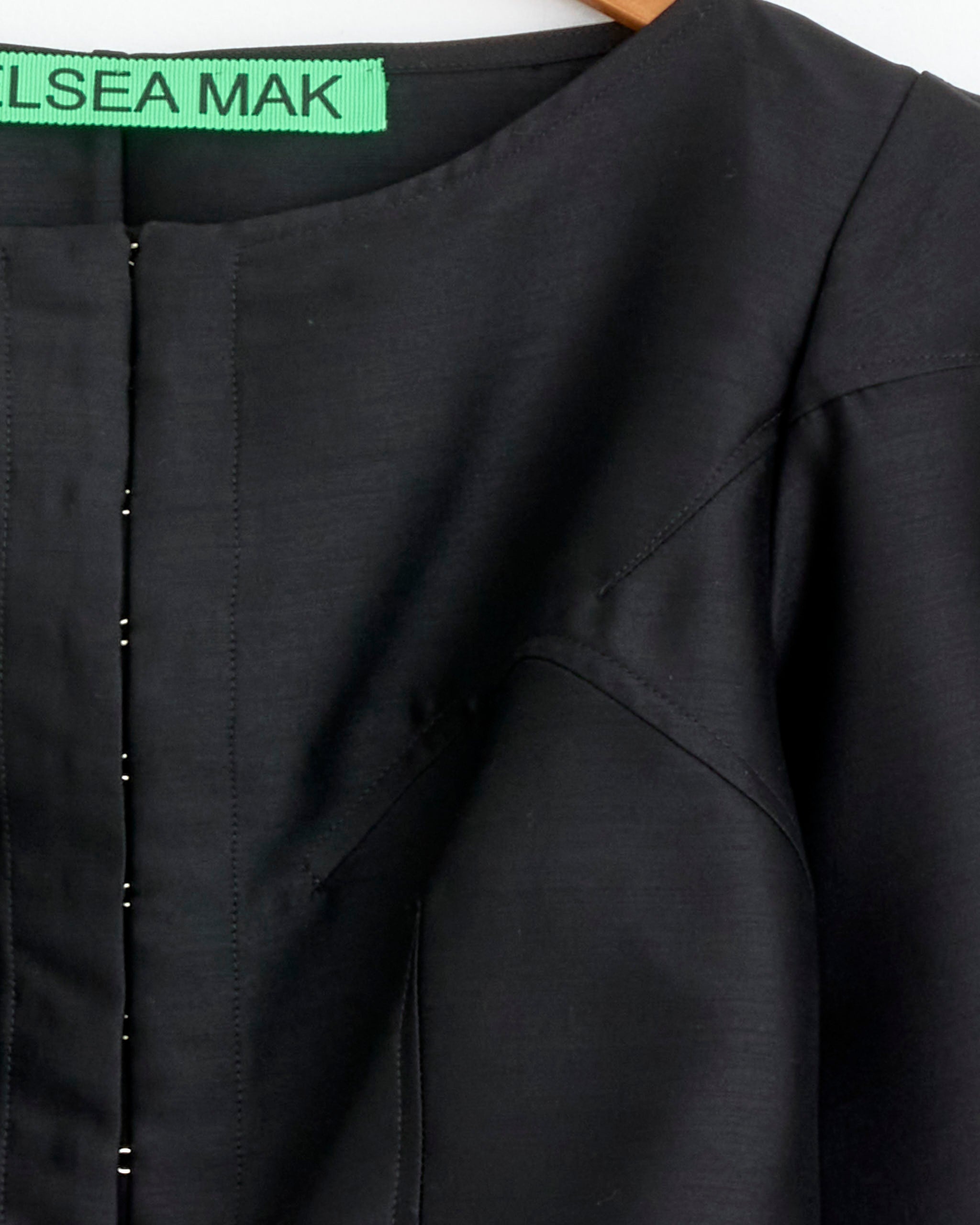 Close-up of the Palace Jacket in Black by Chelsea Mak on a hanger, displaying the upper chest and shoulder with visible seam details and a green Chelsea Mak label.