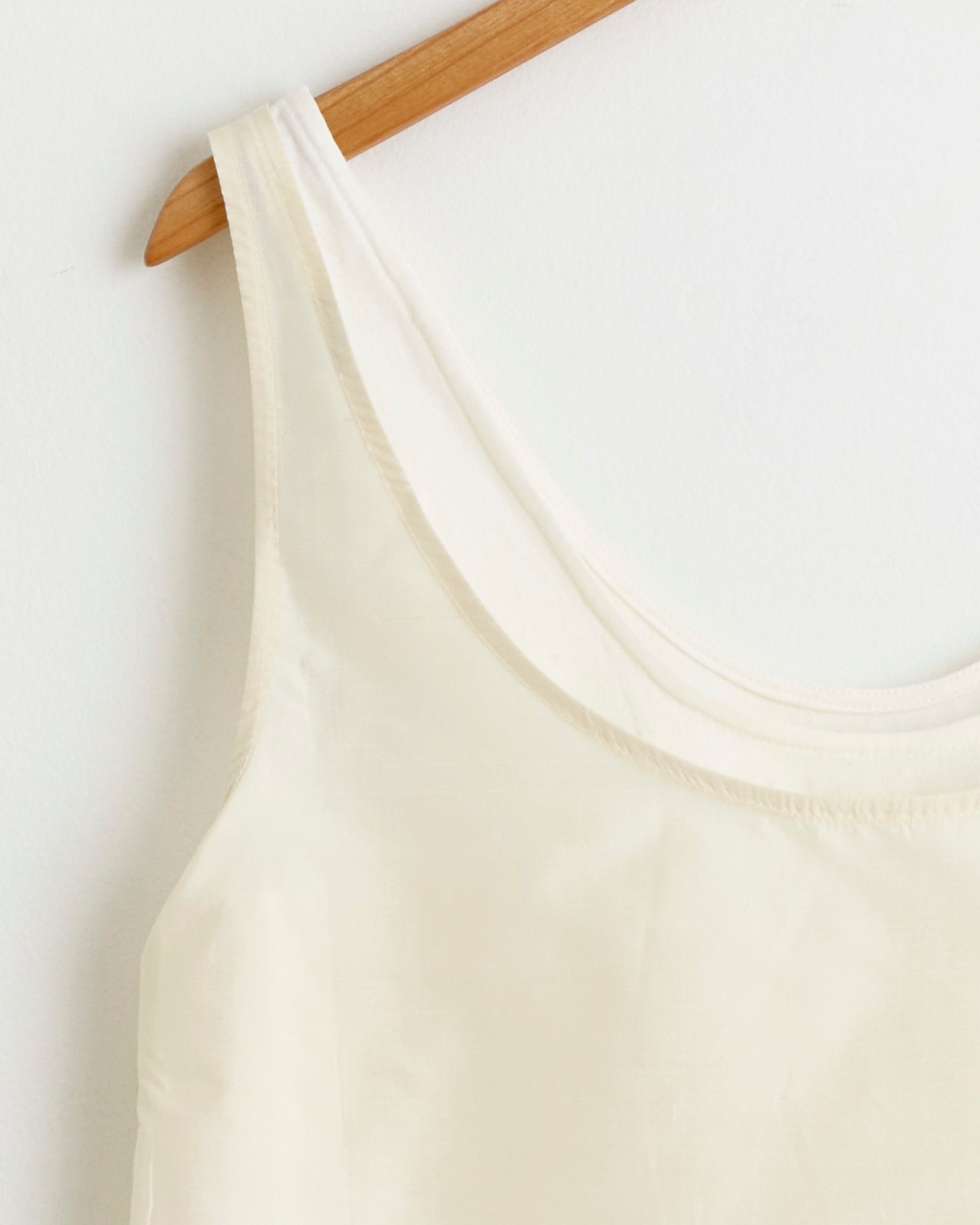 A close-up of the Chelsea Mak Duo Tank in Mixed Whites, a sleeveless modular top, hangs on a wooden hanger against a white wall.