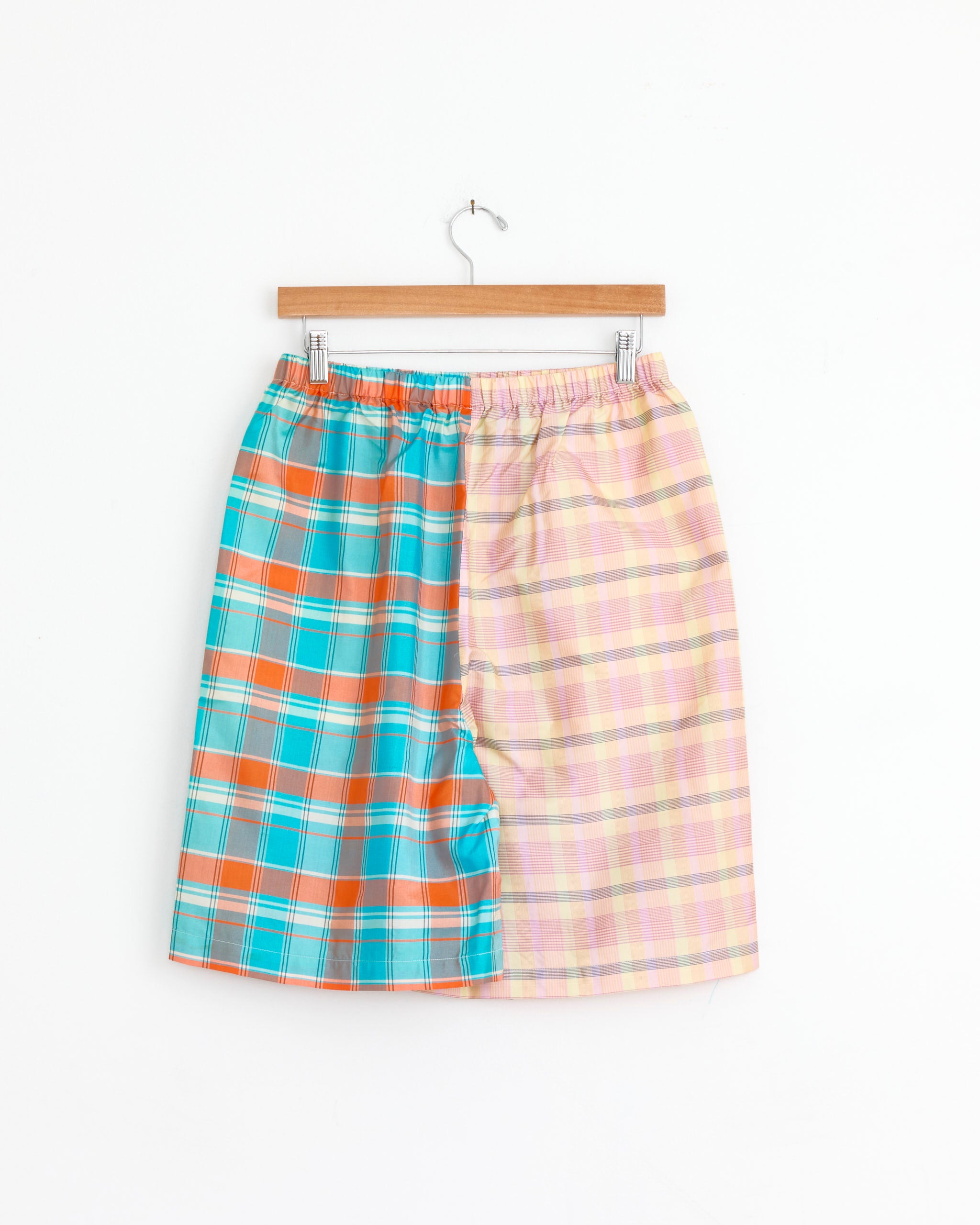 The Chelsea Mak Elio Short in Mixed Plaids features a knee-length cut, elastic waistband, and two contrasting plaid patterns—blue/orange on the left and pastel pink/yellow on the right—displayed on a wooden hanger against a white backdrop.