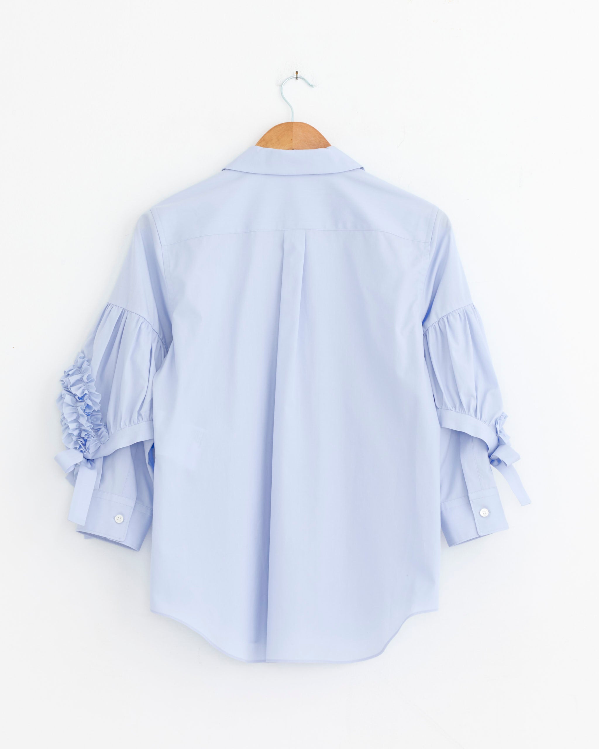 The Comme Des Garçons Girl Ruffled Sleeve Shirt in Blue hangs on a wooden hanger against a white wall, shown from the back. The blouse features ruffled sleeves and ribbon details.
