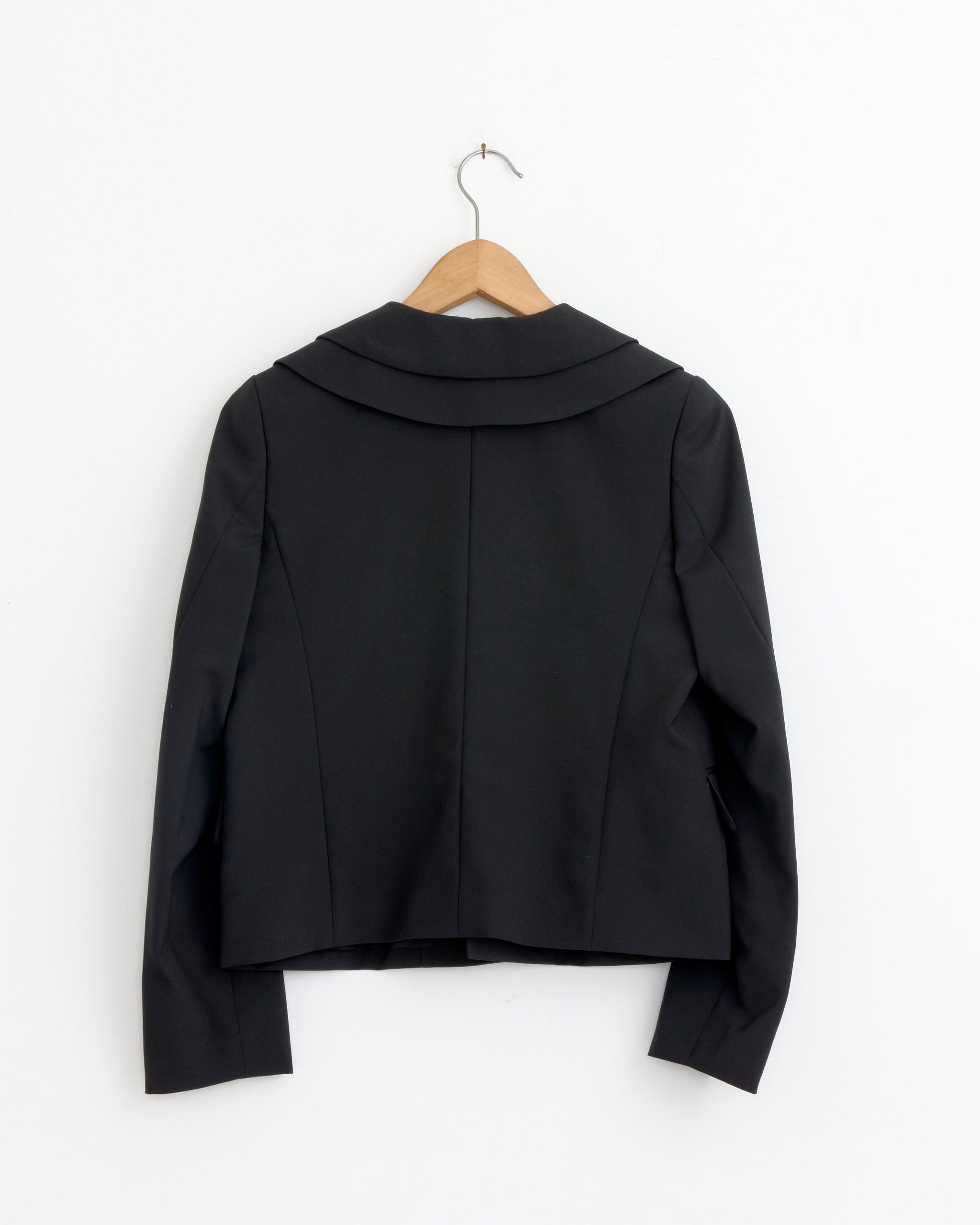 The J015 Jacket in Black by Comme Des Garçons Girl, crafted from 100% wool with a wide collar, hangs on a wooden hanger against a plain white wall, displayed from the back.