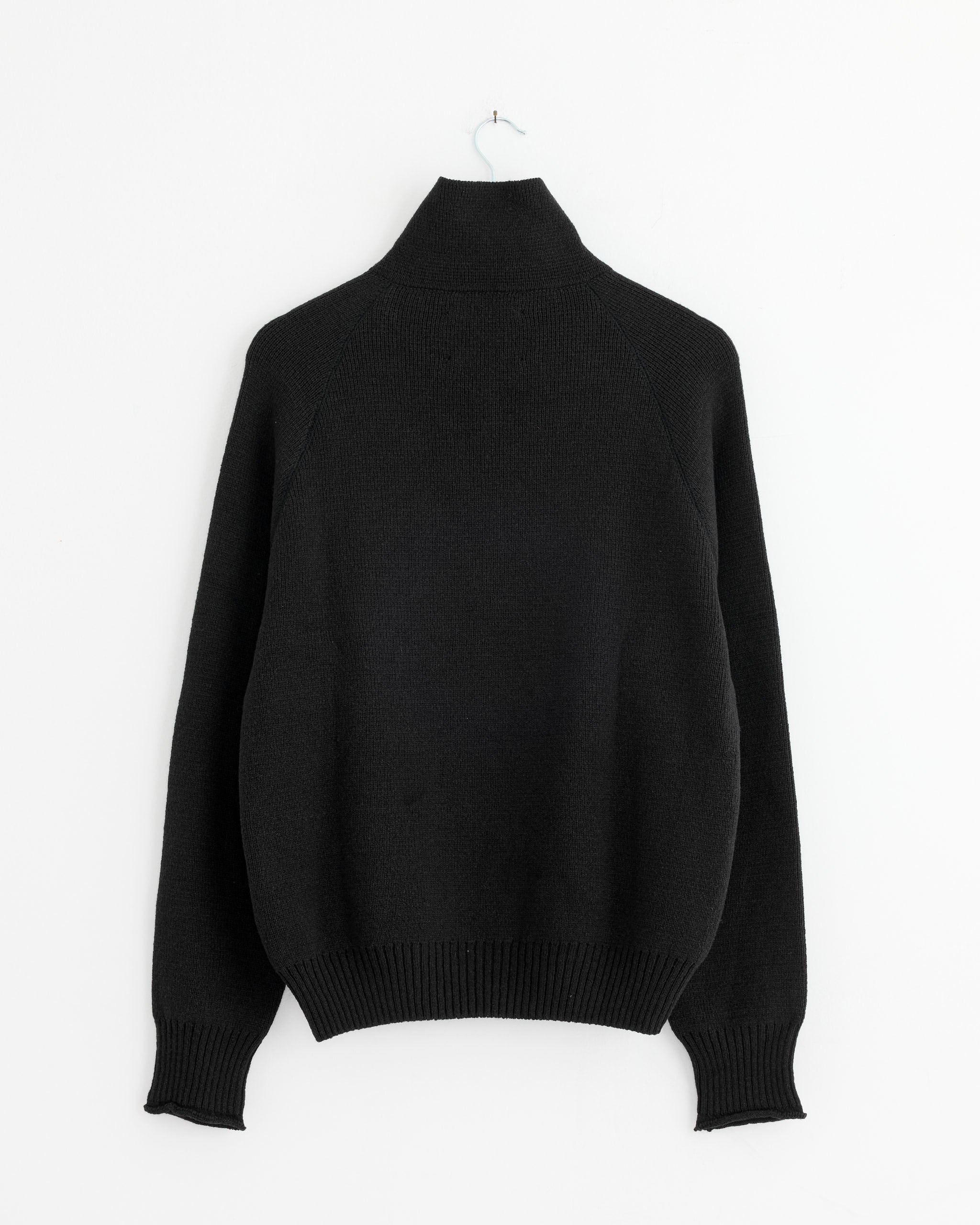 The MHL by Margaret Howell Zip Up Polo in Black, a men's knitwear sweater with long sleeves, hangs on a white wall from a light blue hanger, displayed with the back facing forward.