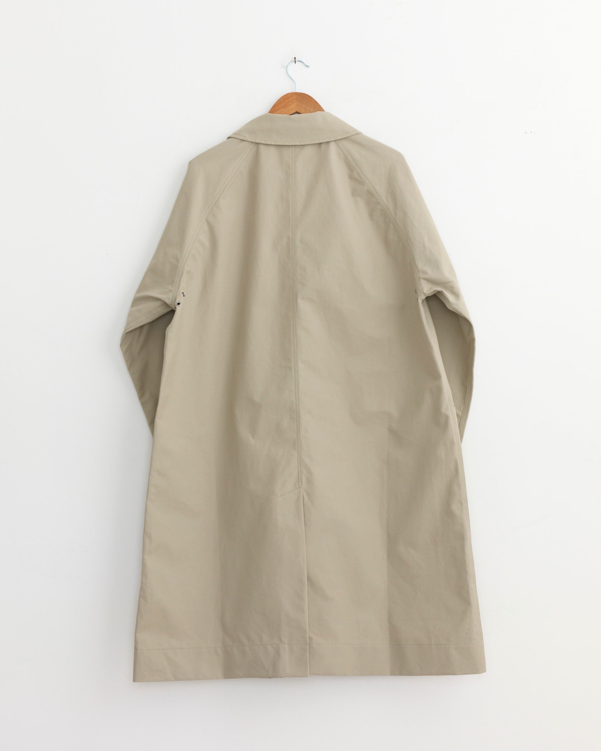 The Railway Mac Trench Coat in Concrete by MHL by Margaret Howell, crafted from showerproof cotton, hangs on a wooden hanger against a plain white wall, displayed from the back.