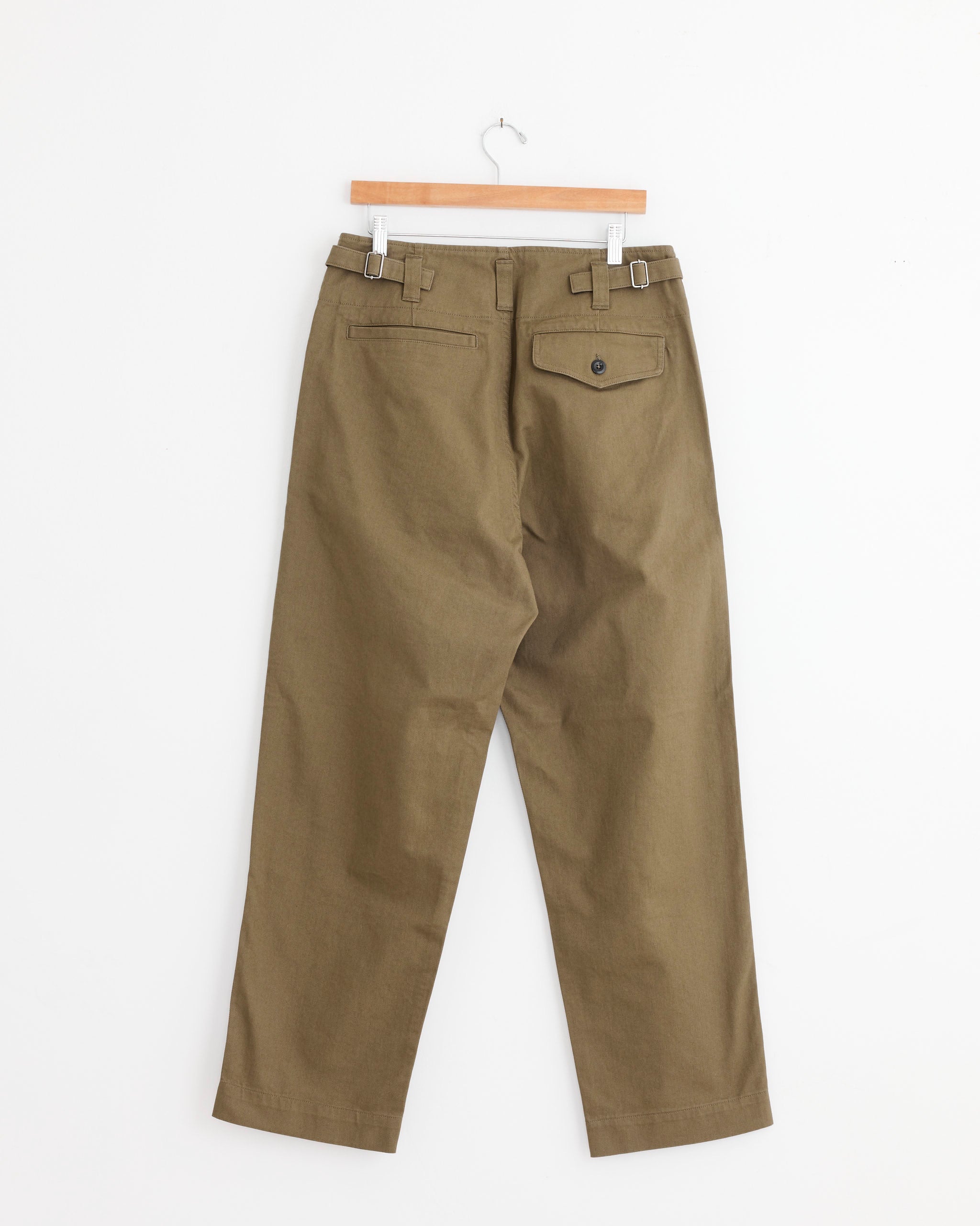 The MHL by Margaret Howell Naval Trouser in Khaki Green, made from cotton drill, hangs on a wooden hanger against a white background, shown from the back with two welt pockets—one with a buttoned flap.
